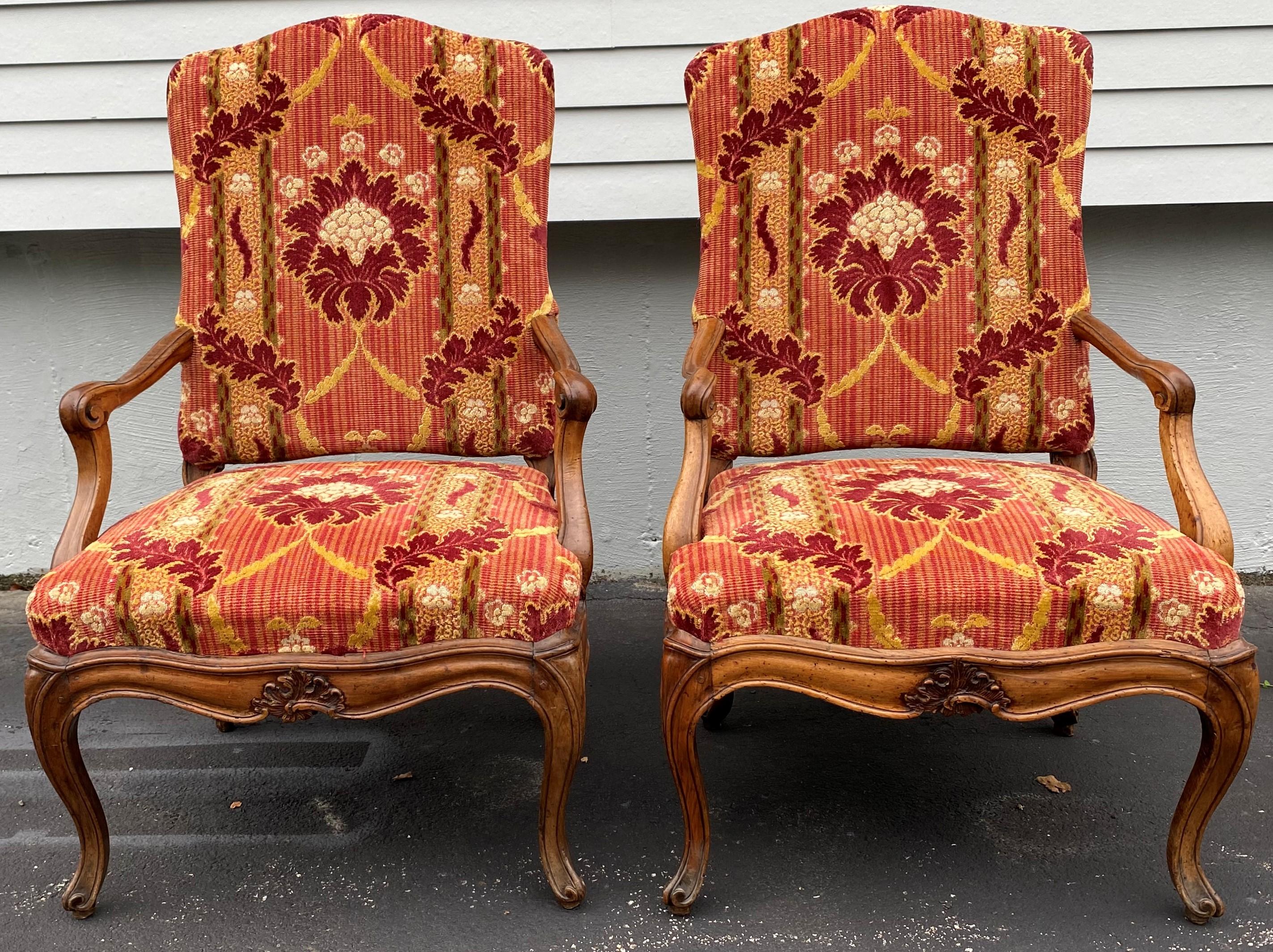 A fine pair of Louis XV style upholstered carved fruitwood armchairs with arched crests, scroll carved arms, a leaf carved center decoration along the front skirt, all supported by four French legs with scroll feet. The backs are finished with a