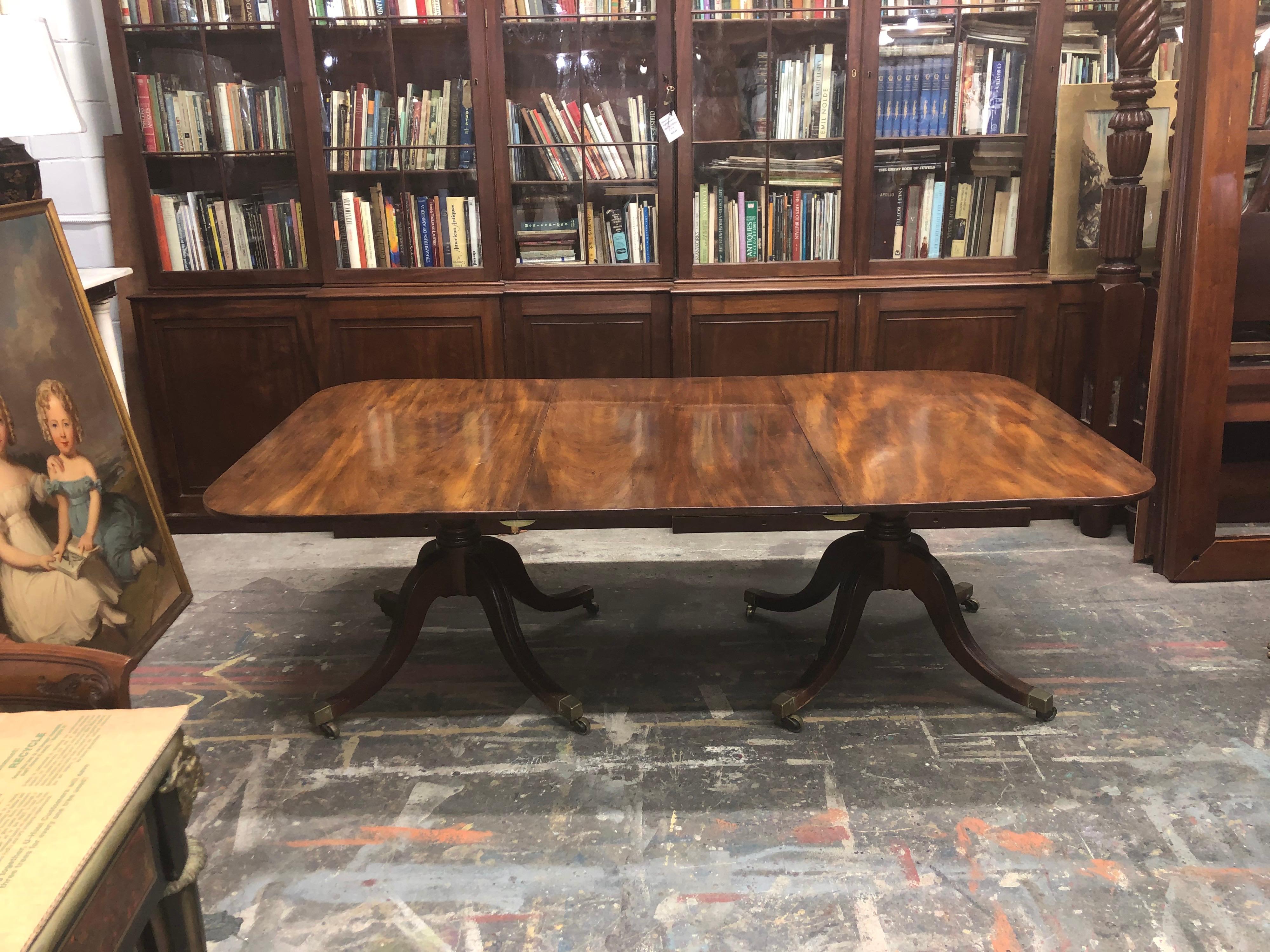 This elegant English Neoclassical double pedestal dining room table has a solid mahogany one board top on each of the pedestal ends and leaf. The original turned pedestals have an urn with rings above and are supported by four mahogany double carved