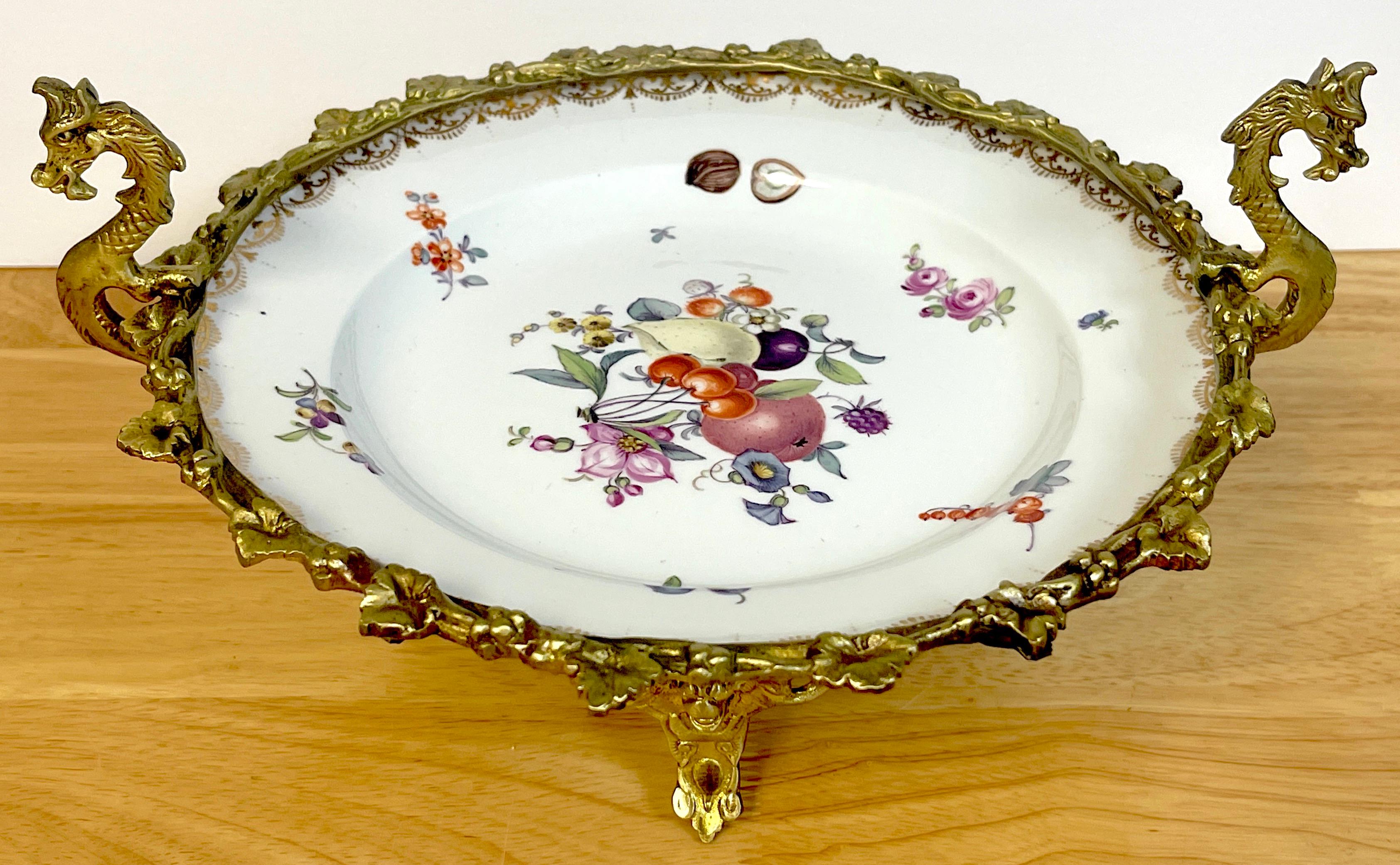 19th C Meissen Fruit & Flower Motif Ormolu Mounted Tazza, The Meissen porcelain (possibly late 18th century) finely decorated with various fruits, nuts, and flowers. Held in a twin handled griffin motif grape encrusted footed ormolu frame.