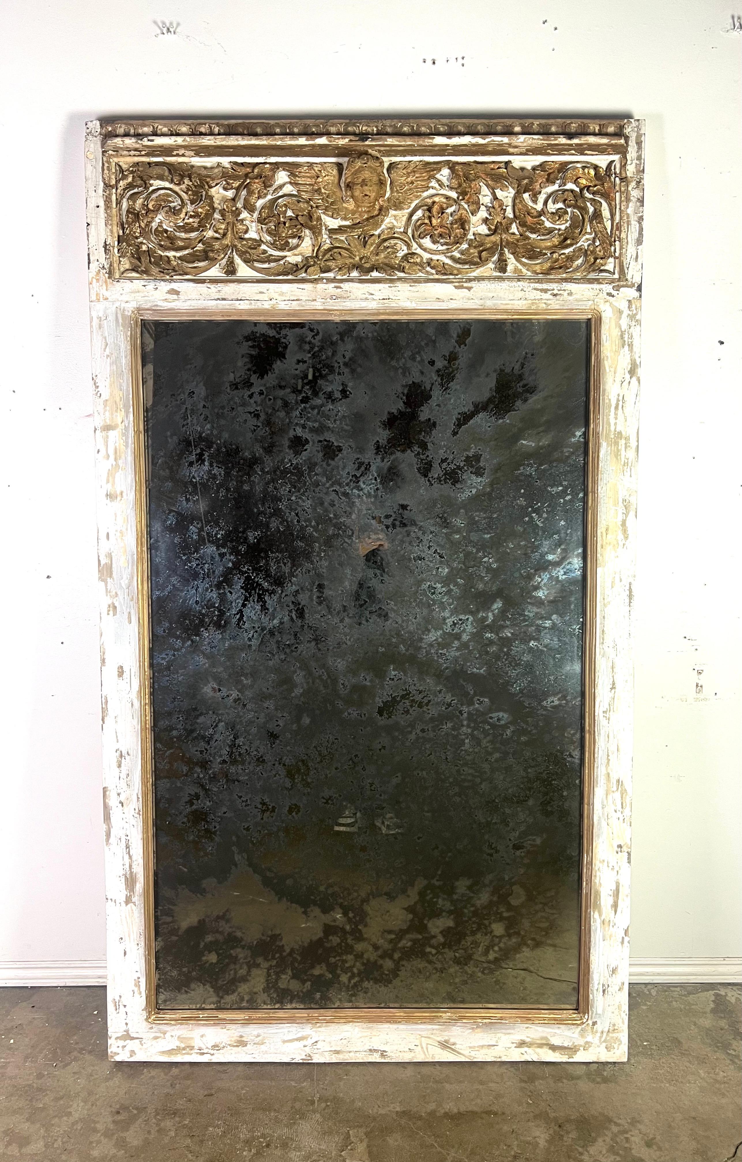 Specchio italiano del XIX secolo dipinto e dorato a parcella.  La cornice dello specchio presenta intagli ornamentali nella parte superiore, tra cui motivi floreali e a volute con una figura centrale simile a un cherubino.  La cornice è dipinta di