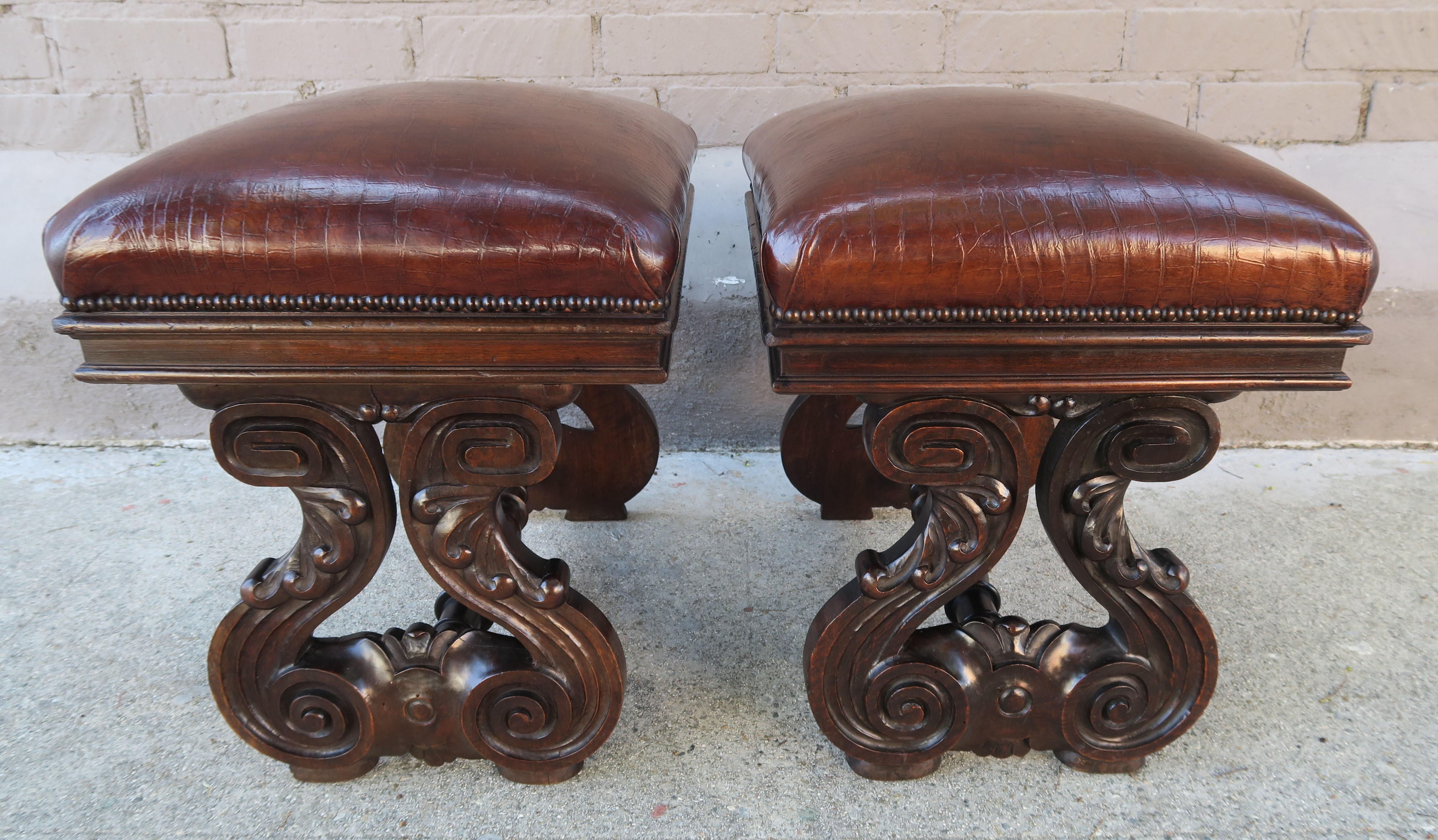 Pair of 19th century Italian carved walnut benches that are upholstered in a beautiful worn embossed tobacco colored leather with antique brass nailhead trim detail. The benches stand on two scrolled legs with carved acanthus leaves connected by a