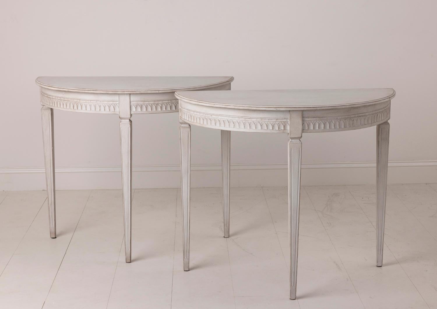 A petite pair of Swedish Gustavian Style demilune console tables from the 19th century. The tables feature lamb's tongue carving on the apron; raised on square, tapered, and fluted legs. These demilune tables would make beautiful hall or bedside