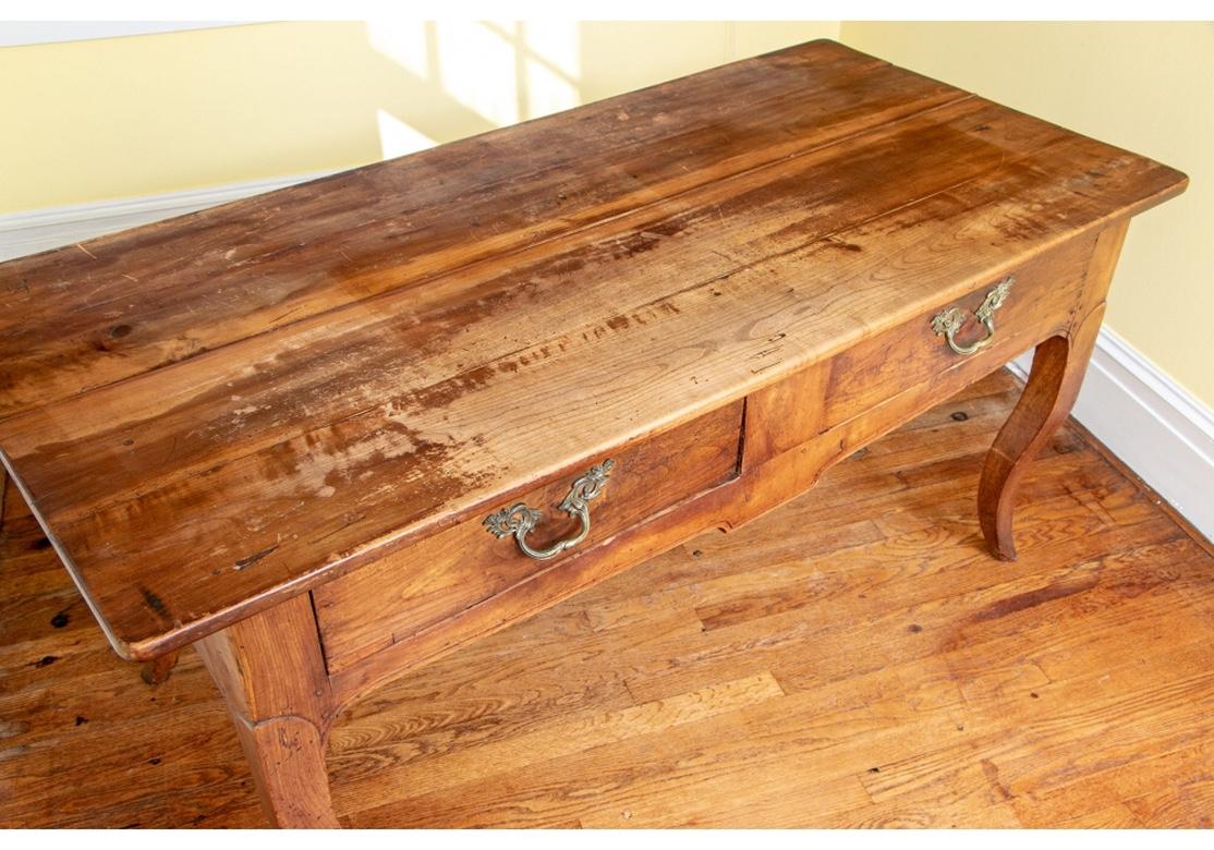 A long plank top overhanging with rounded corners. The shaped apron on one side with leafy vine and flower pot inlays. The other side with two drawers (cracks) with baroque style brass pulls. Shaped side aprons. Raised on angular tall tapering
