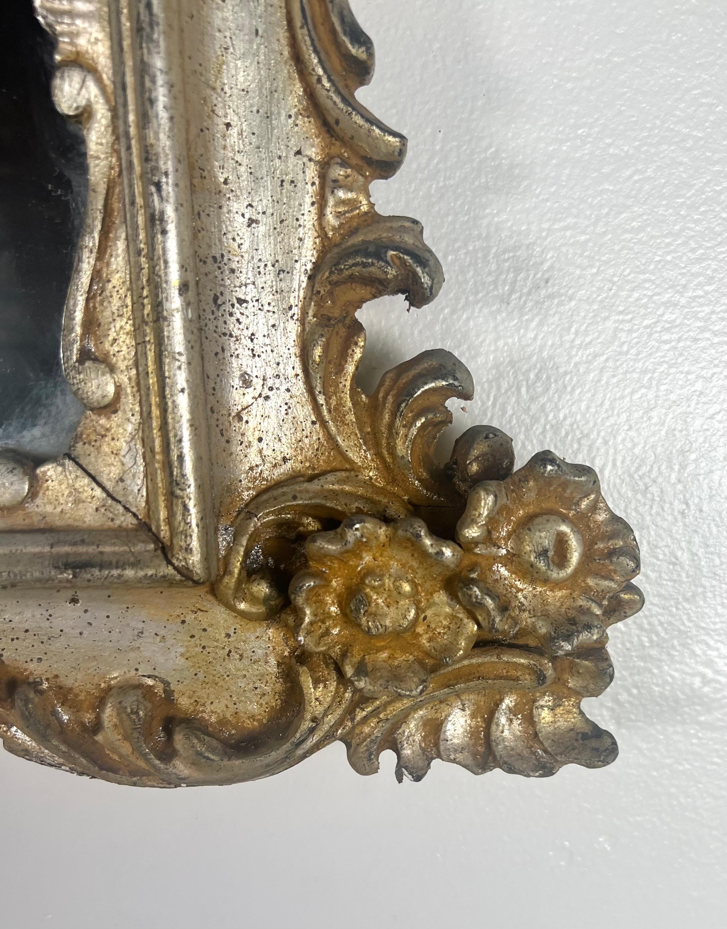 The silvered gilt wood mirror with gold highlights on the carvings is inspired by Rococo styles.  Its elaborate gilt wood frame features an array of intricate carvings, including acanthus leaves, florals, and scrolled elements, which are typical of