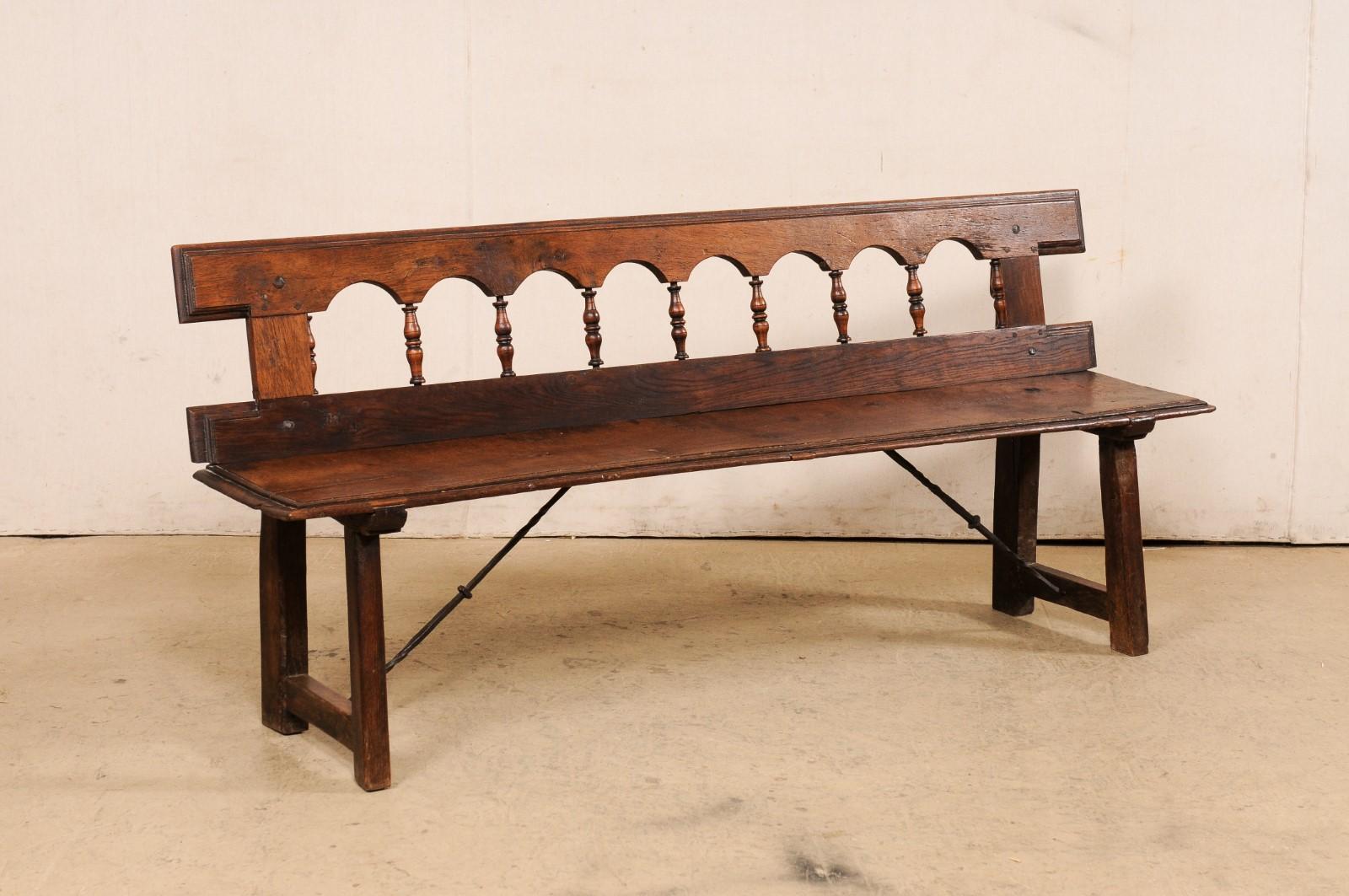 A Spanish carved-wood bench, with trestle-legs and iron stretchers, from the 19th century. This antique wooden bench from Spain features a back rest carved in a repeating open arch motif with spindle back-splats and straight-edge top rail. The sides