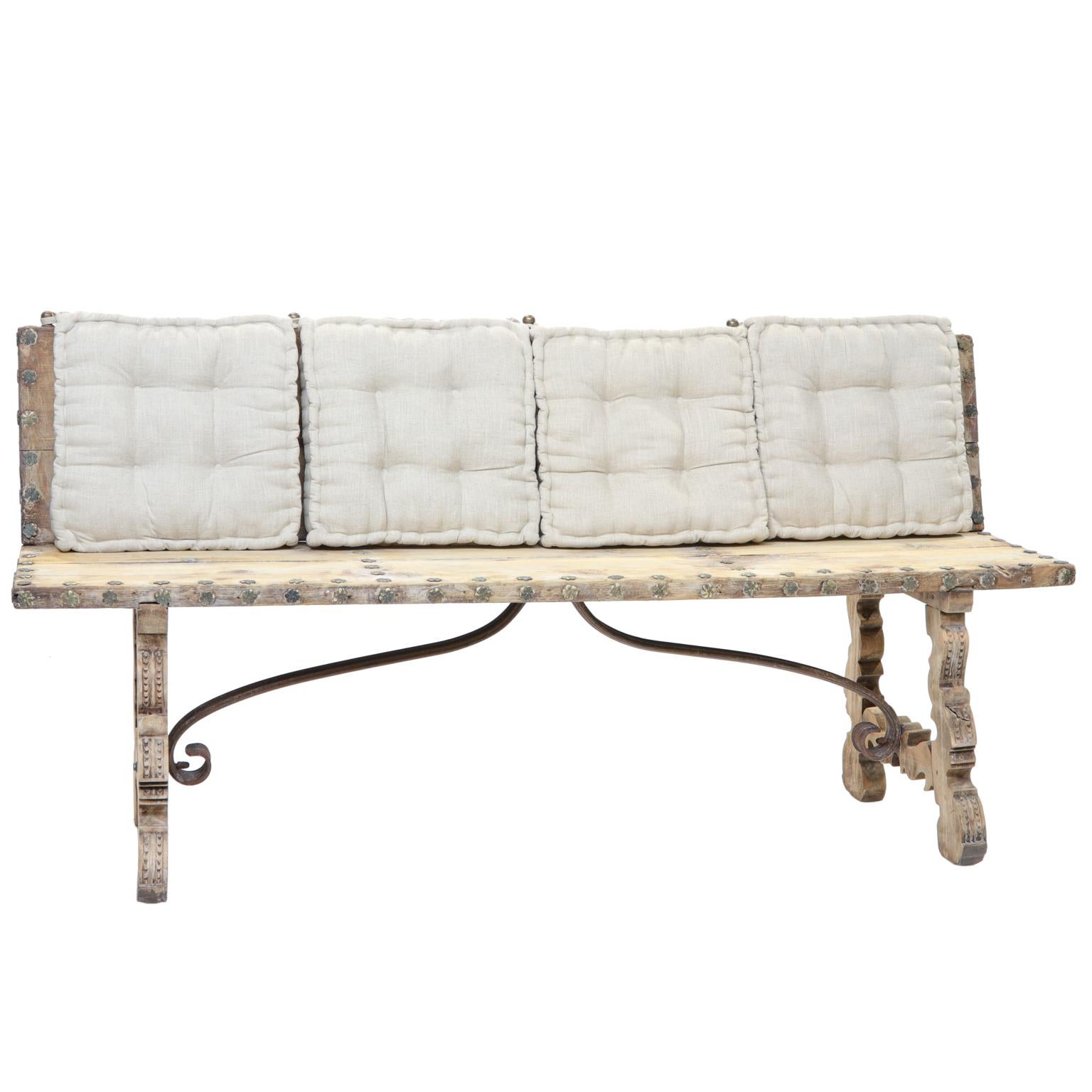 A 19th century Spanish bench. This bench was most likely covered at one point. It has been taken down to the frame and stripped and bleached. Nailheads are old and give this a great look. Carvings adorn the legs and are connected by an iron