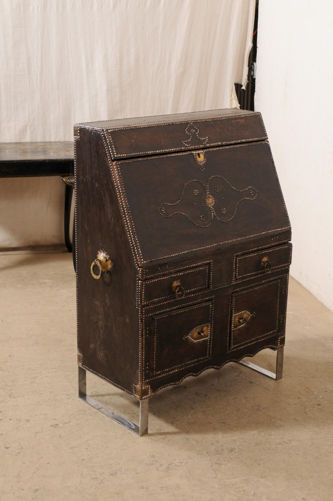 A 19th century Spanish secretary desk, on custom iron base. This antique case piece from Spain has a leather-wrapped exterior which is adorn in nail-head accents, original iron hardware, and nicely scalloped skirt at front. When the fall-front is