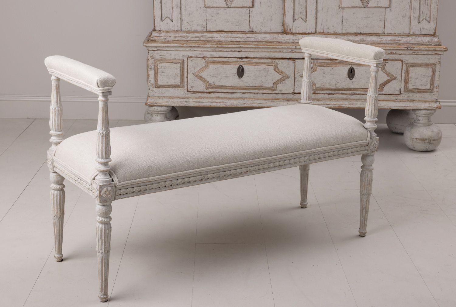 A rare Swedish window bench in the Gustavian style newly upholstered in fine Belgian linen with padded armrests. The frame features beautifully carved acanthus leaves and bell flowers with rosettes on the corner posts ending in tapered and fluted