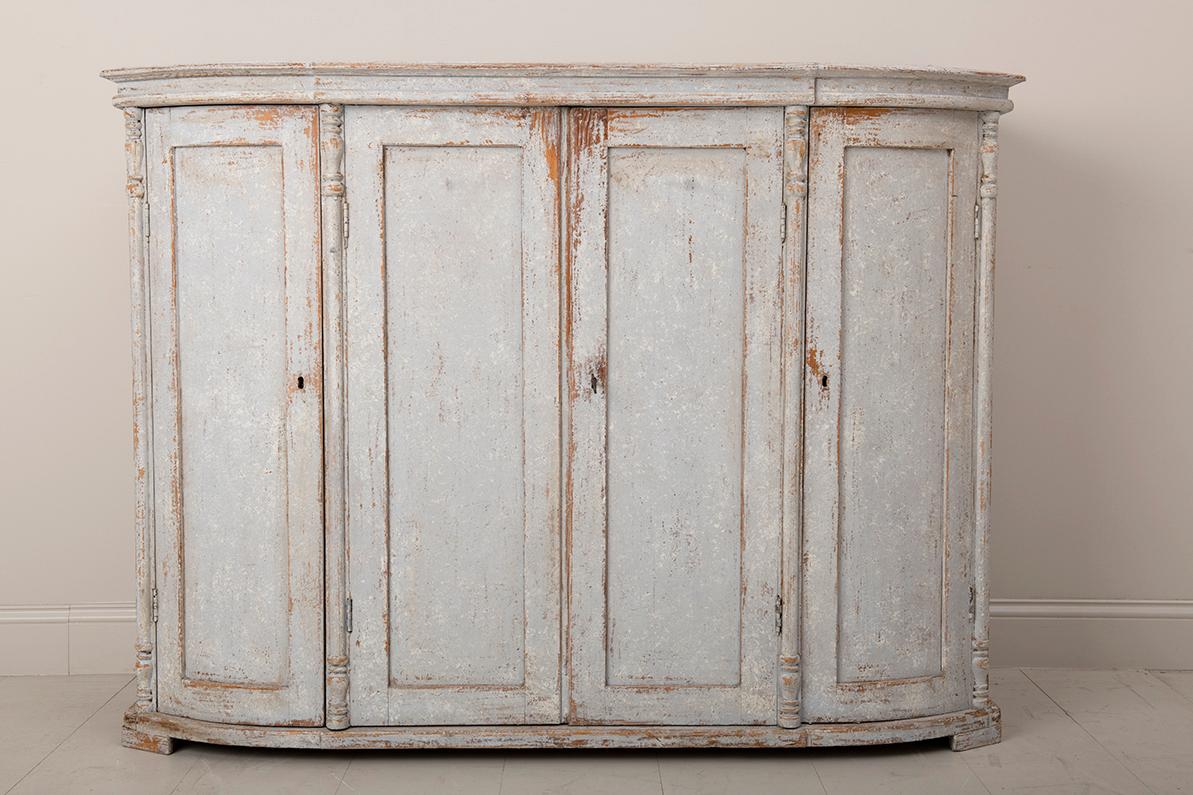 A Swedish demilune sideboard or server from the Gustavian period. The patina is a beautiful, chalky, neutral ivory with a slight gray hue and natural wood showing through in areas. There are three interior shelves behind four doors.  The center