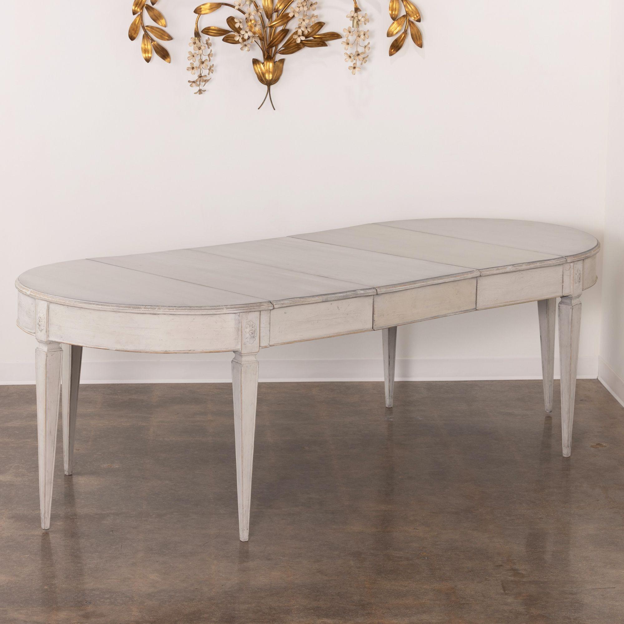 A beautiful, Swedish, three-leaf, oval dining table in the Gustavian style with a light gray patina. There is a carved rosette above each square, tapered, and fluted leg. This is a charming and versatile table that can extend with one, two, or three