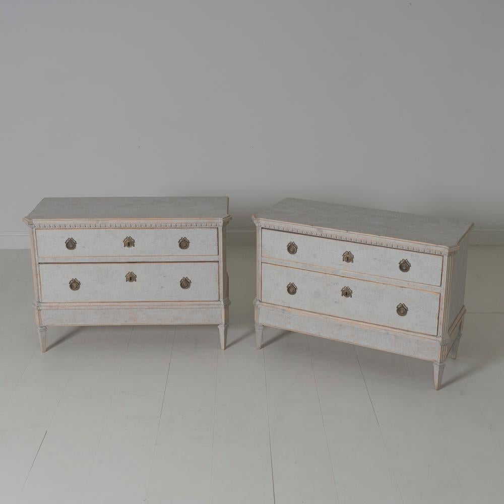 A pair of Swedish commodes in the Gustavian style with dentil molding around the top. Chalky antique ivory paint. Two large drawers, canted corner posts, and tapered legs. Two keys.