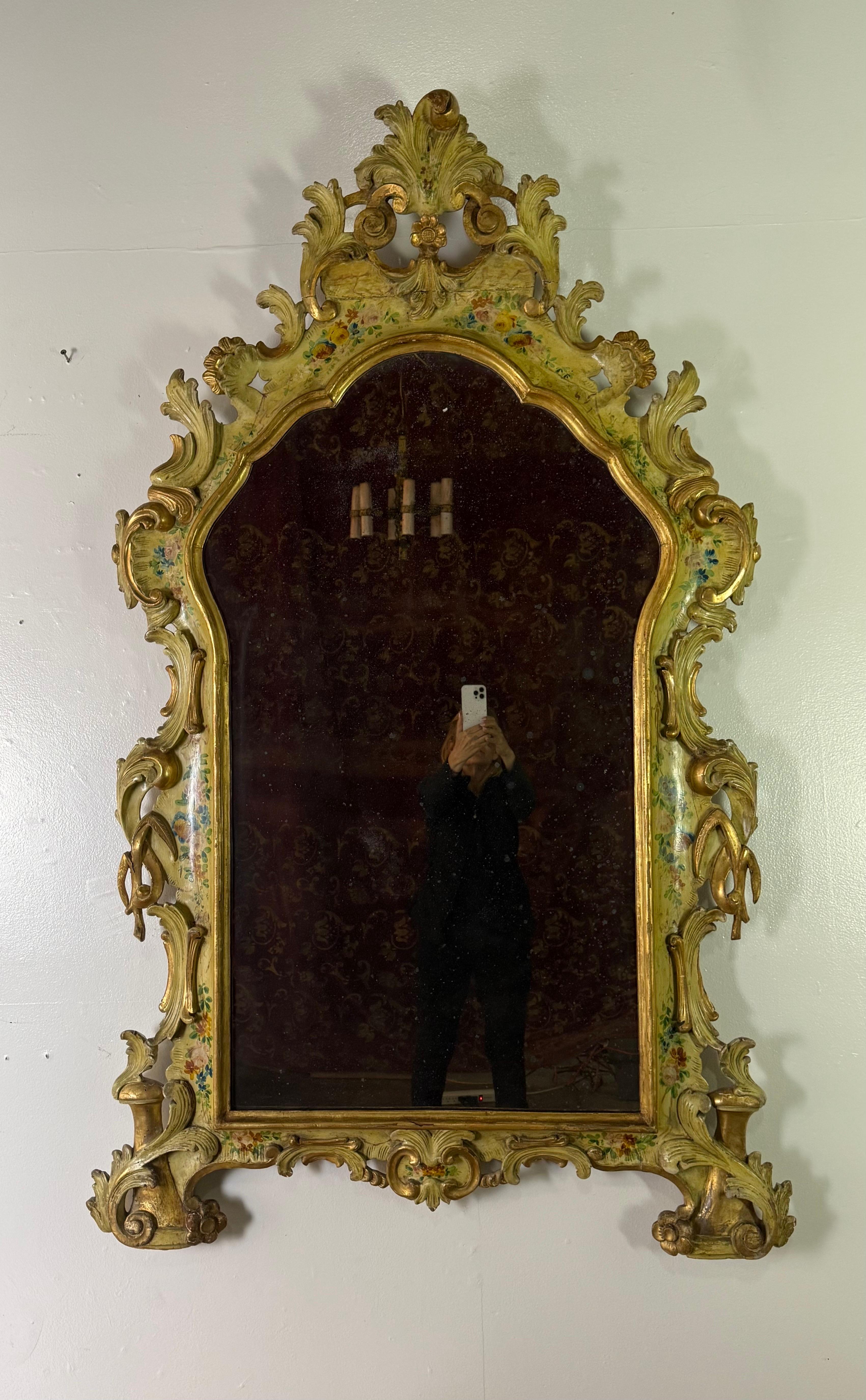 19th C. Venetian mirror with a heavily carved and hand-painted frame. The wood is richly detailed with scrolls, acanthus leaves, and floral motifs, all contributing to a dramatic silhouette. The surface of the frame is painted in a soft, aged