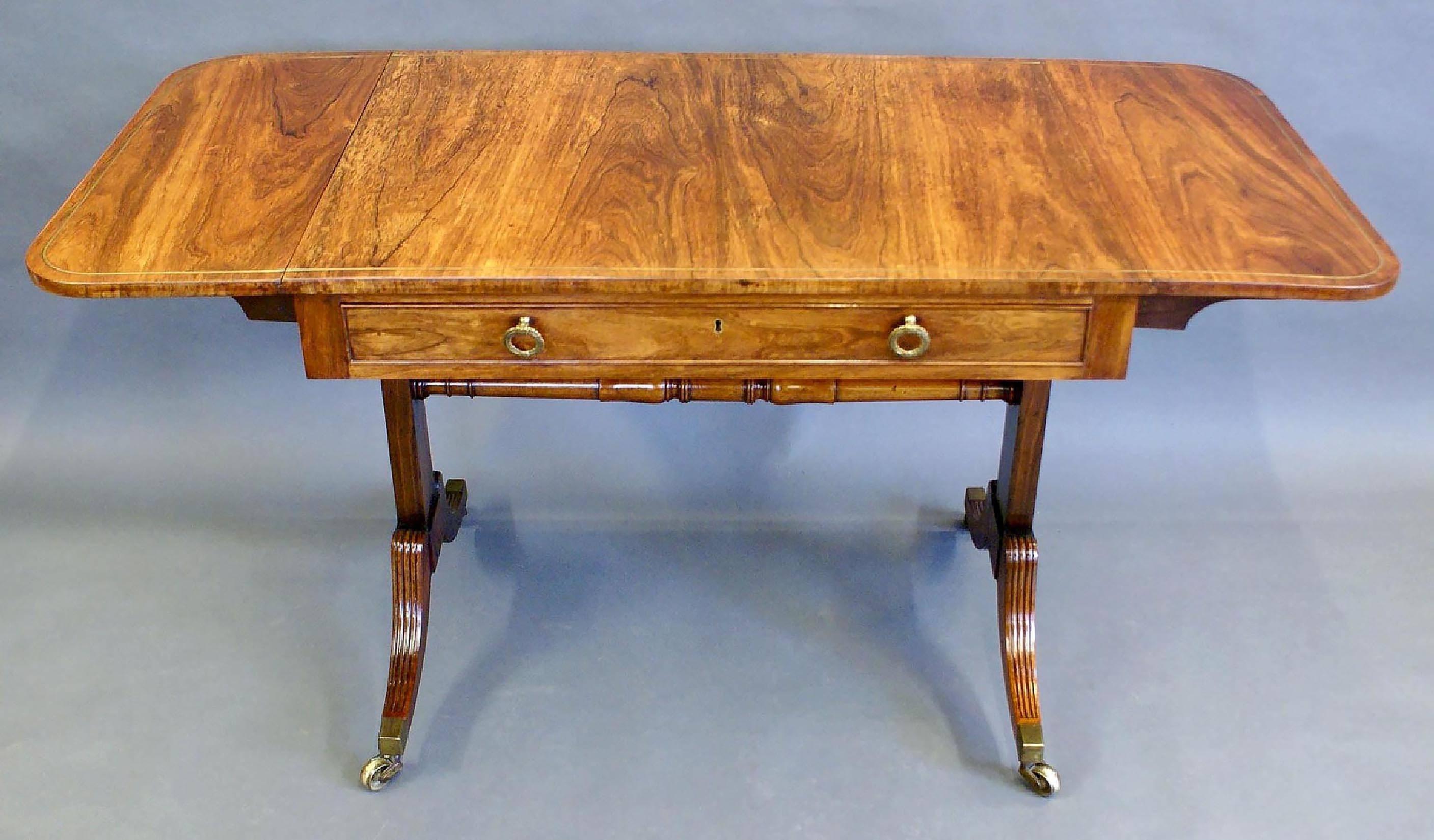 This beautiful and excellent quality English rosewood sofa table features one top drawer with a matching false drawer on the opposite side, and a brass stringing border trim. The table is supported on reeded Sabre legs and stretcher base, with all