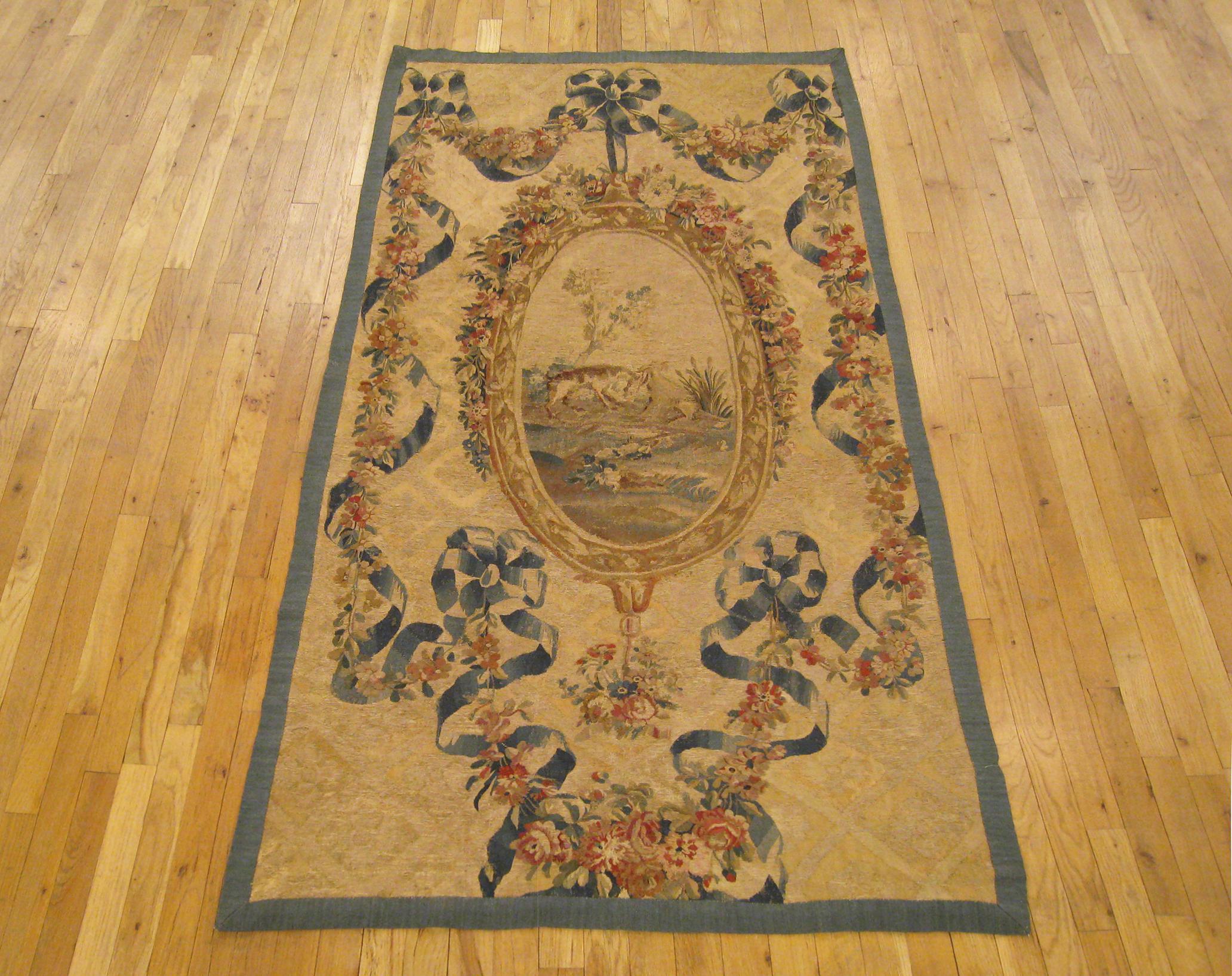 An antique French Aubusson needlepoint tapestry from the 19th century, featuring an oval pendant at center, which envisions an idyllic landscape. The pendant medallion is surrounded by floral garlands and ribbon ties, with soft color tones in the