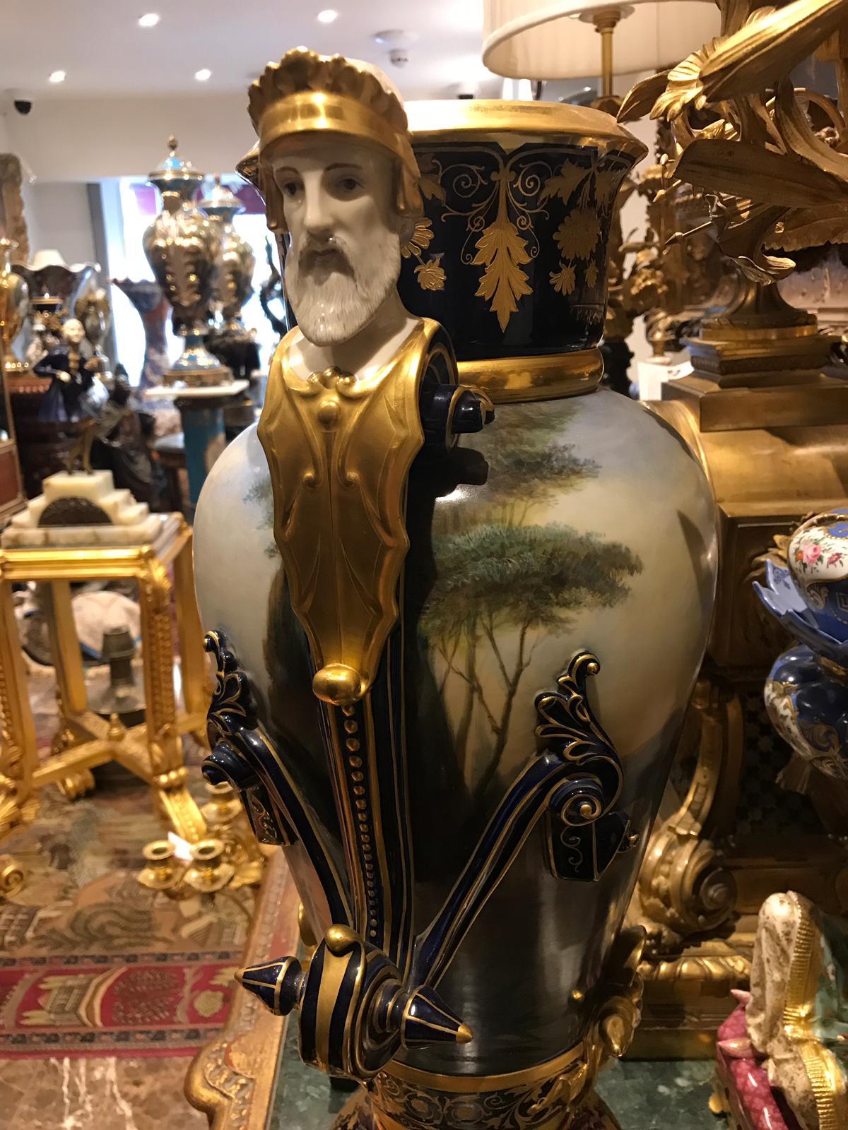 Of a baluster form, the pair feature scroll handles topped by a neoclassical crowned male's bust (mythological bust) and another face around the base portion above the foot. The dark blue vases are painted with Neapolitan peasant girls against