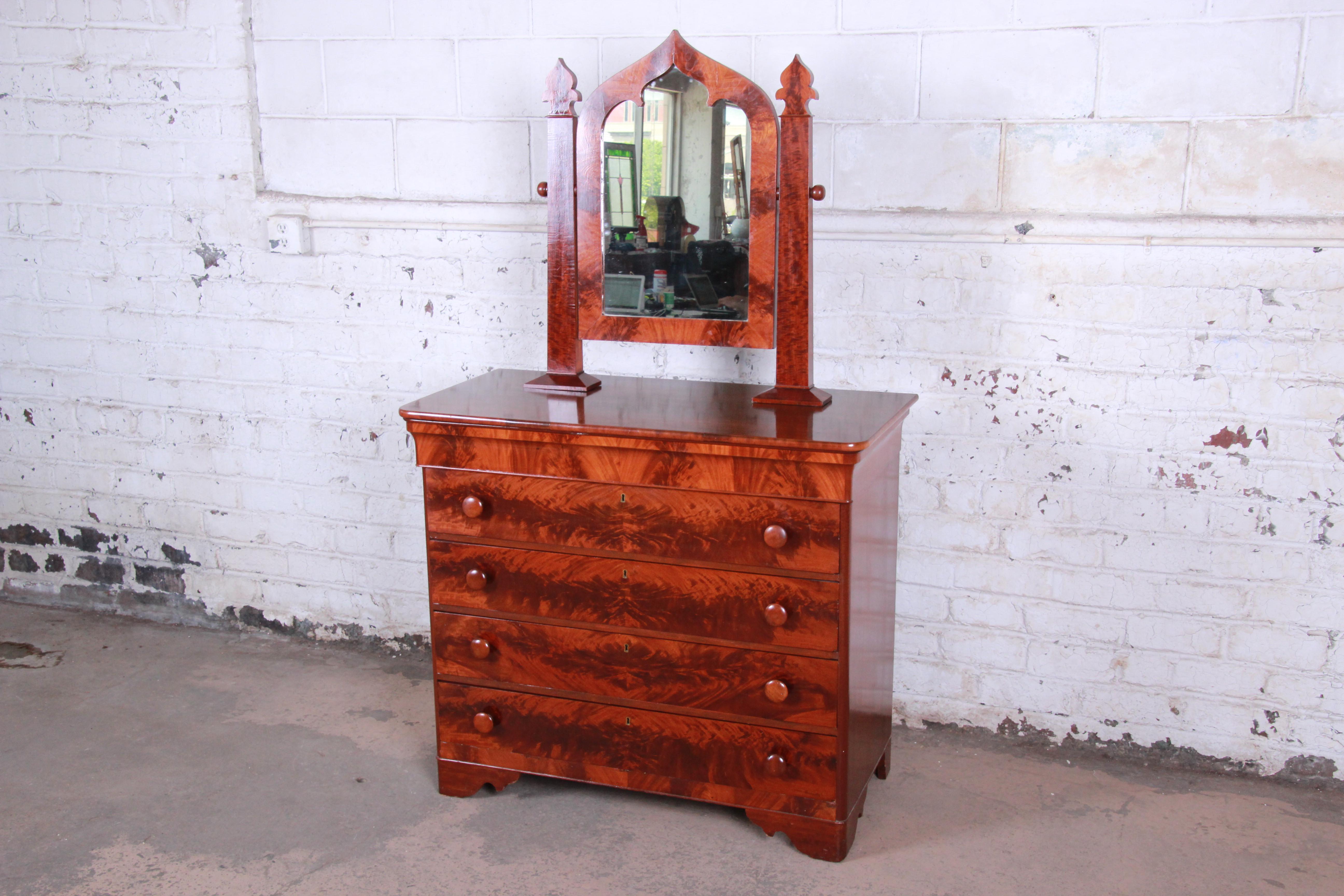 An exceptional 19th century American Empire mahogany dresser with mirror. The dresser features stunning flame mahogany wood grain and a unique Gothic Cathedral style swing mirror. It offers ample storage, with five dovetailed drawers. The dresser is