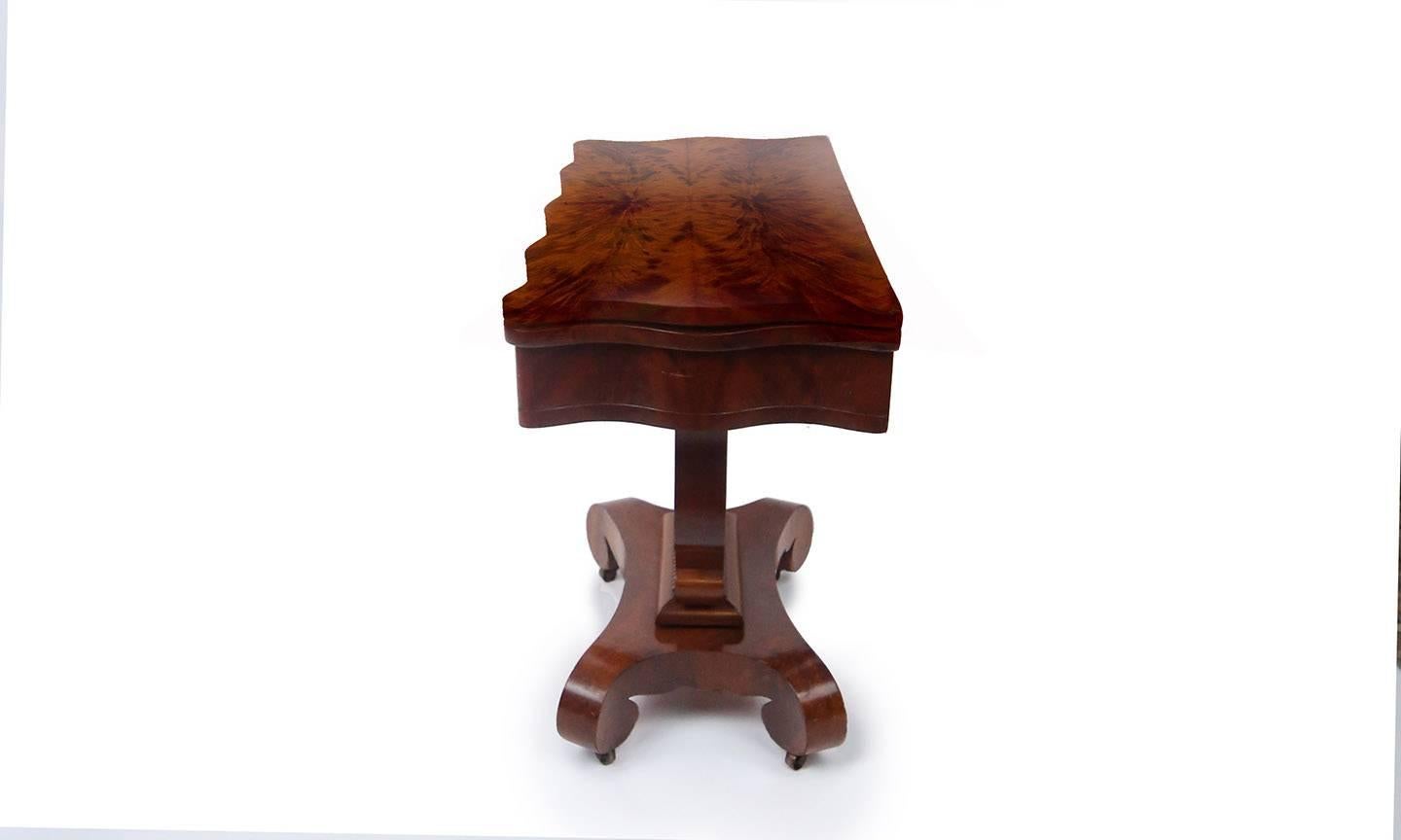 Handsome Empire card or tea table with centre pedestal in mahogany and matchbook flame mahogany. Hinged top opens to reveal compartment and pivots to become square table. Scalloped edge top and frieze supported by bold harp base and scroll legs with