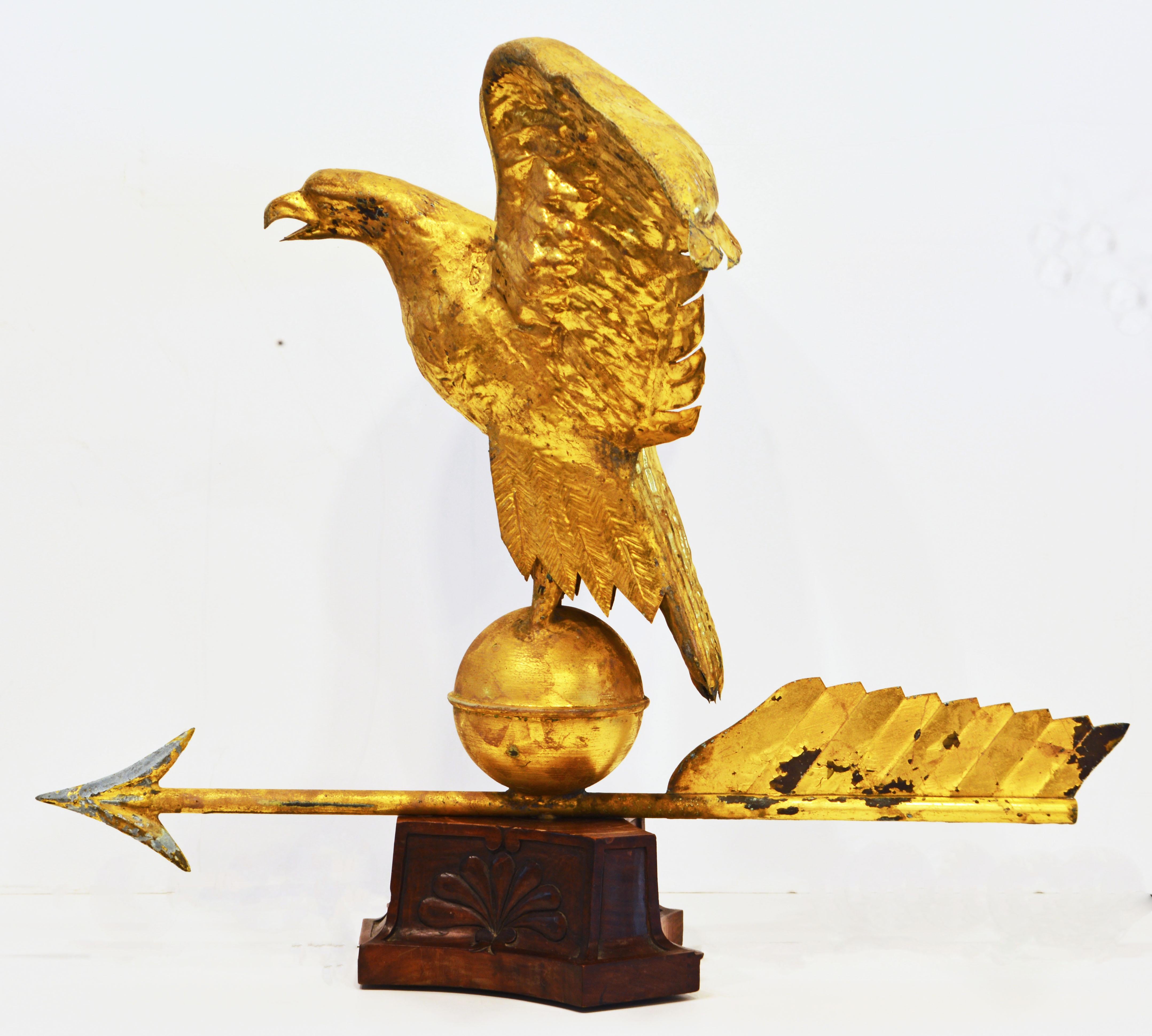 With wings spread high and richly molded detail this gilt on copper eagle weathervane, dating to the late 19th century, is mounted on a belted gilt ball on an arrow resting on a carved triangular wood base.