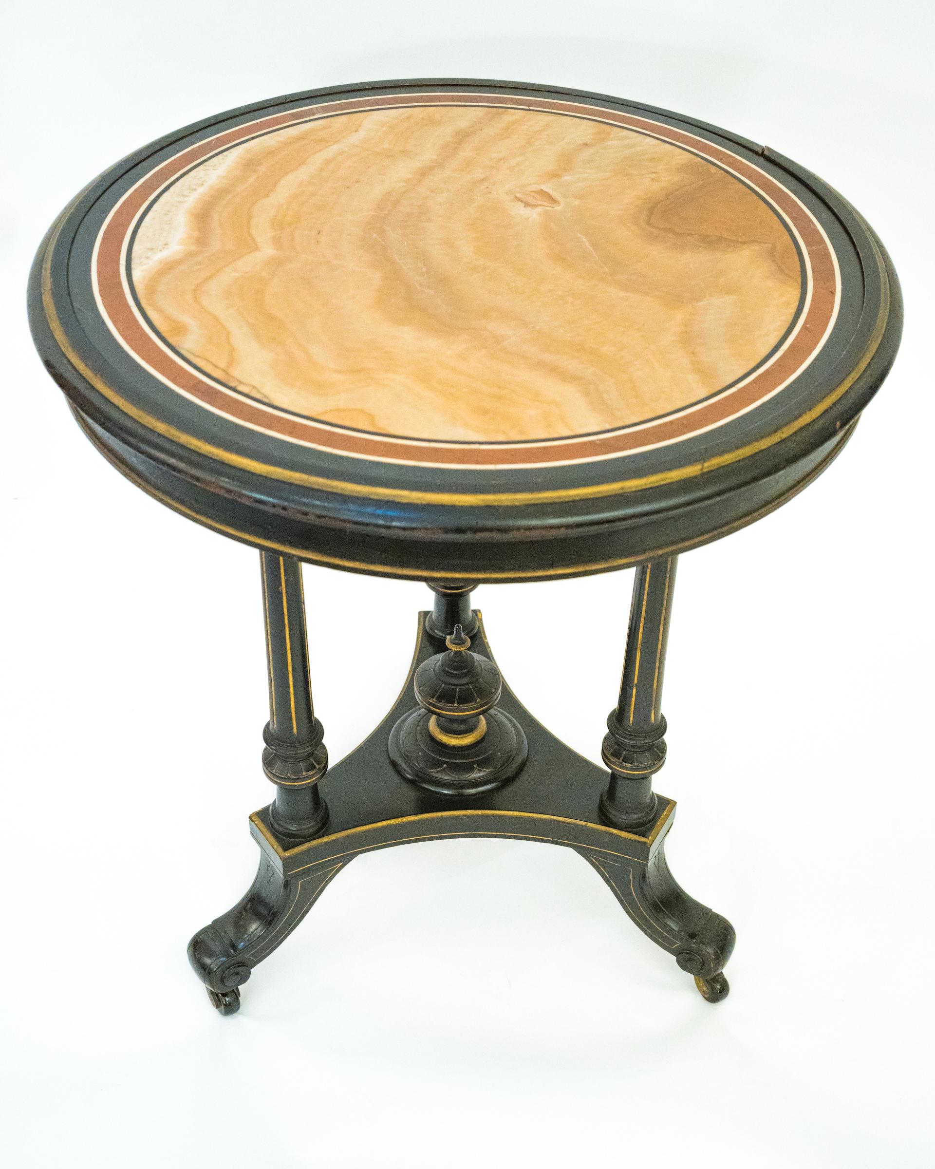 An American Greek Revival side table with 3 tapered fluted round legs supported by triangular plinth supported by splayed legs and having center finial. The ebonized frame with gilt details and specimen onyx top with contrasting red, cream, and