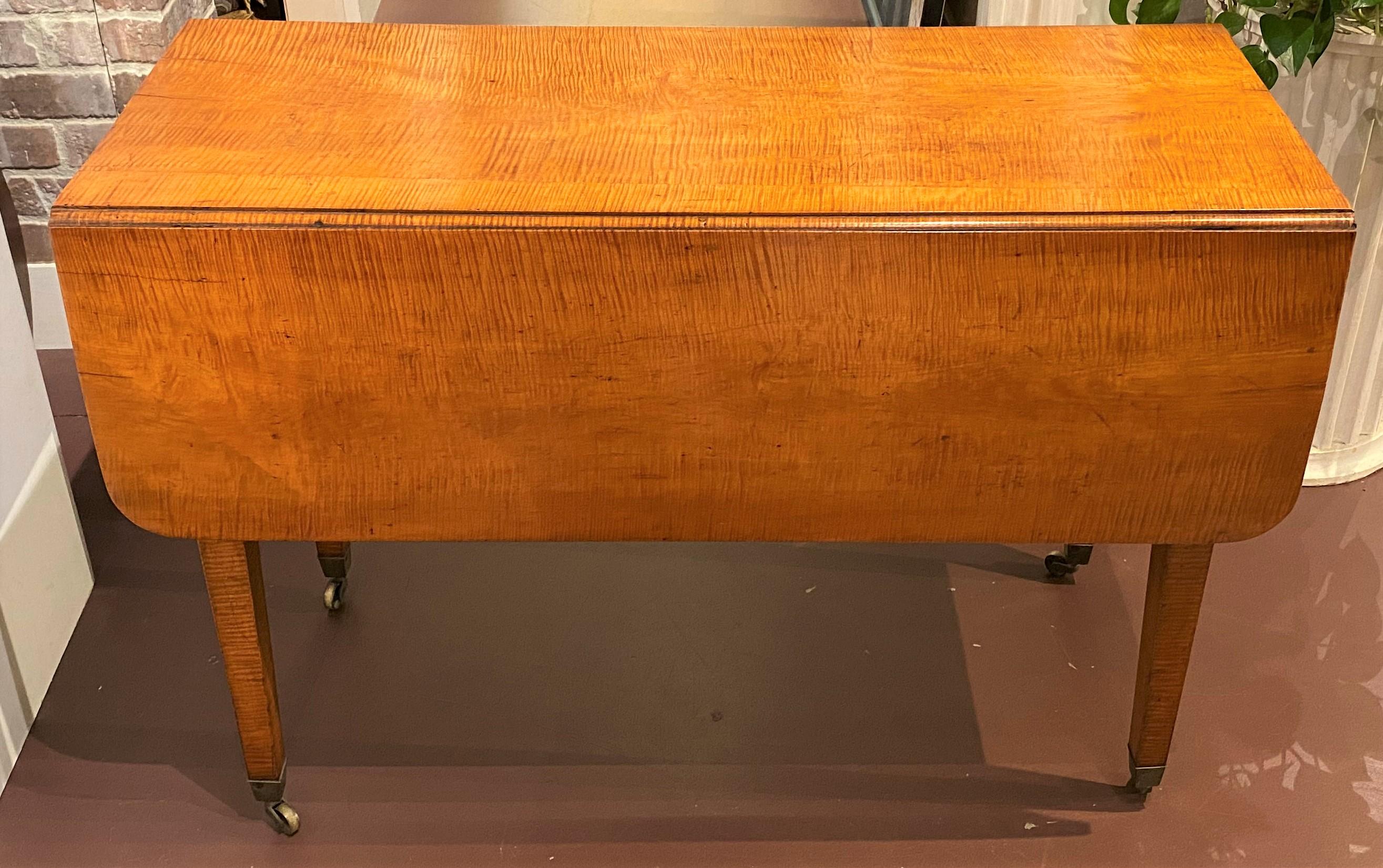A fine example of an American Hepplewhite tiger maple rectangular drop leaf table with rounded corners and four square tapered legs, two of which being gate legs, all terminating with brass cap casters. With leaves open, the table shows off its