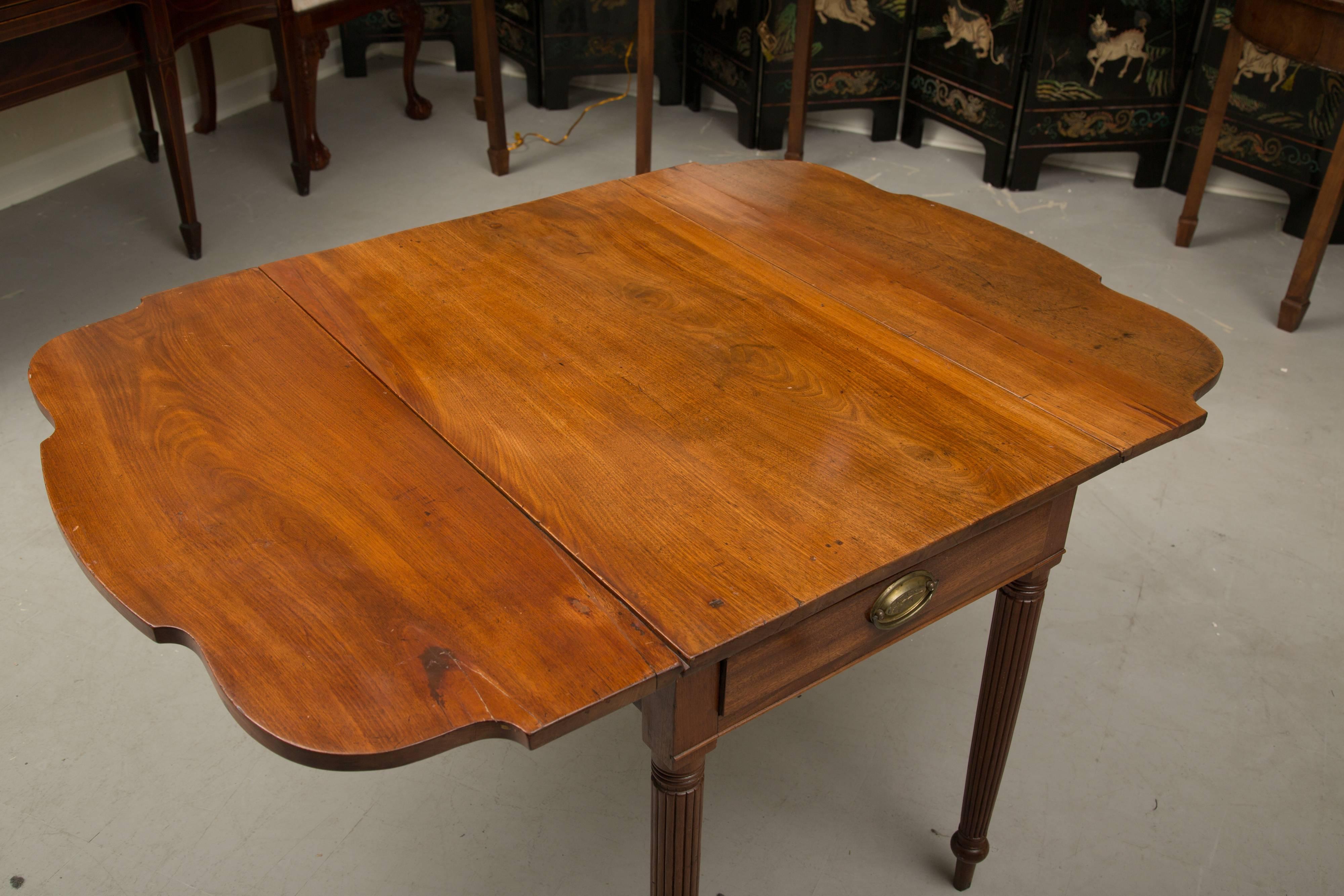 This is a mellow American Sheraton pembroke table. The top is flanked by scalloped drop leaves above a frieze drawer and raised on circular reeded legs, early 19th century.