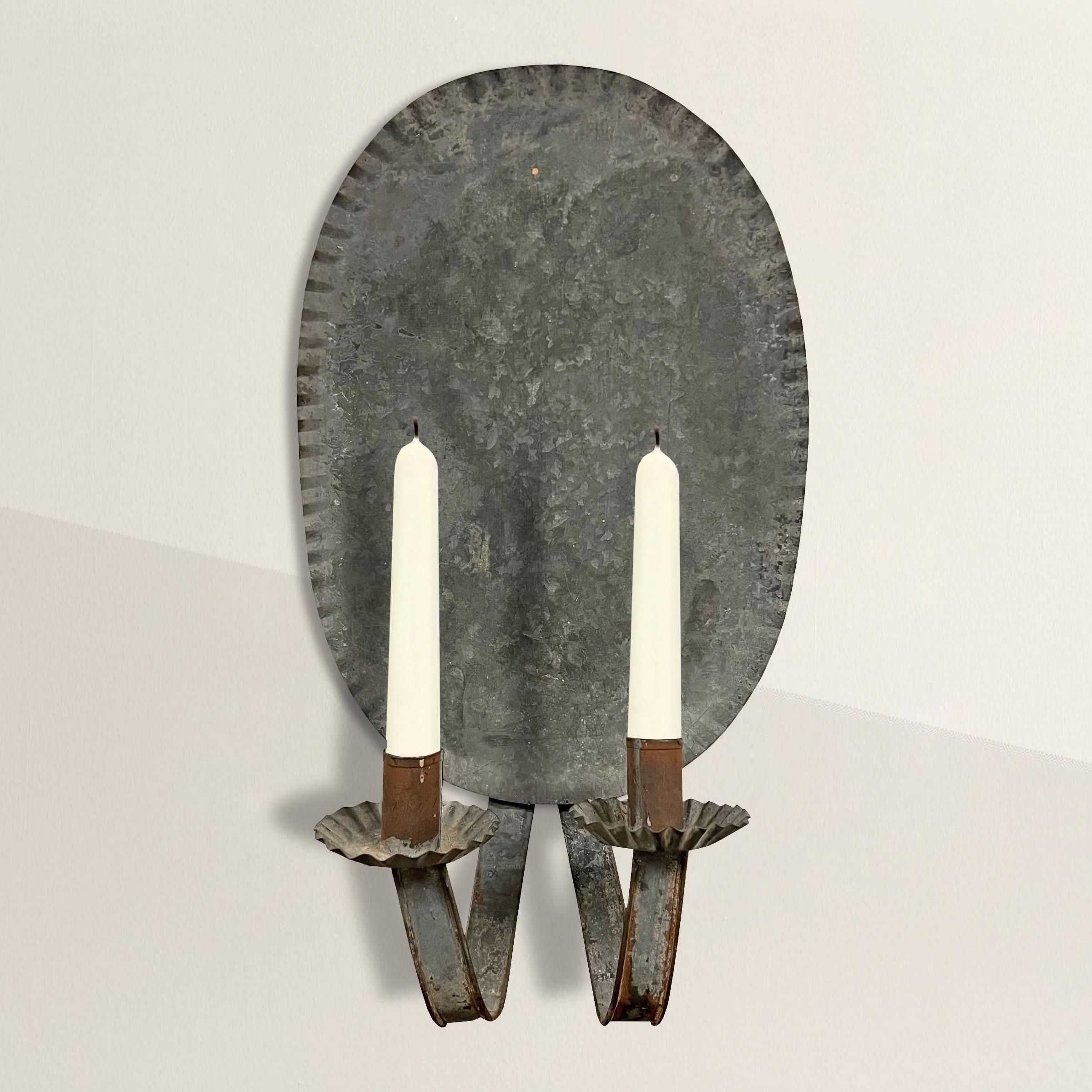 A chic 19th century American two arm tin candle sconce with crimped oval backplate and candle cups. A beautiful piece to hang somewhere you need a little extra sparkle at night.