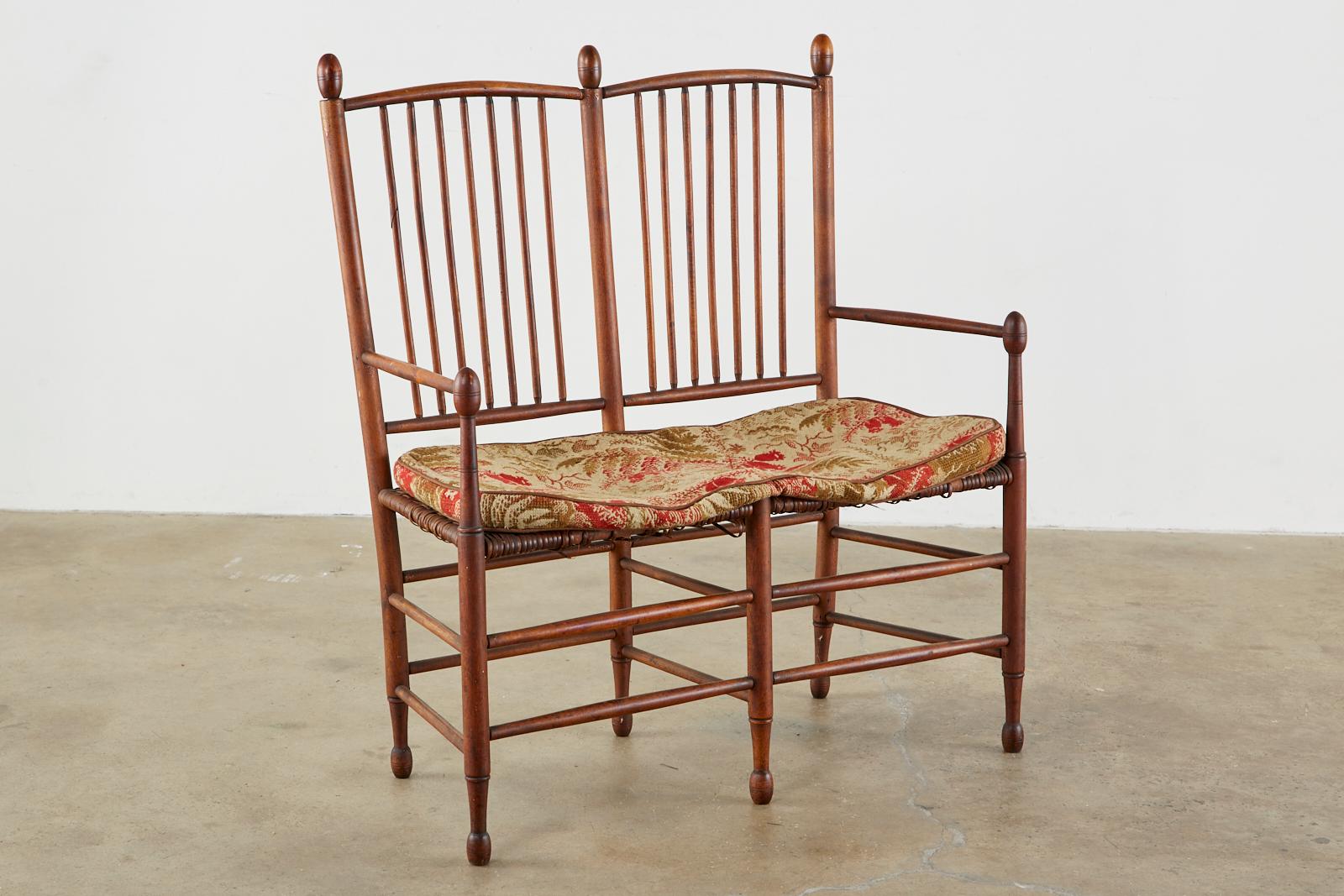 Rustic 19th century American Windsor settee or bench constructed from maple with a rush seat featuring a spindle back. The arms and back are topped with round finials that match the feet. The seat shows signs of wear with an embroidered seat