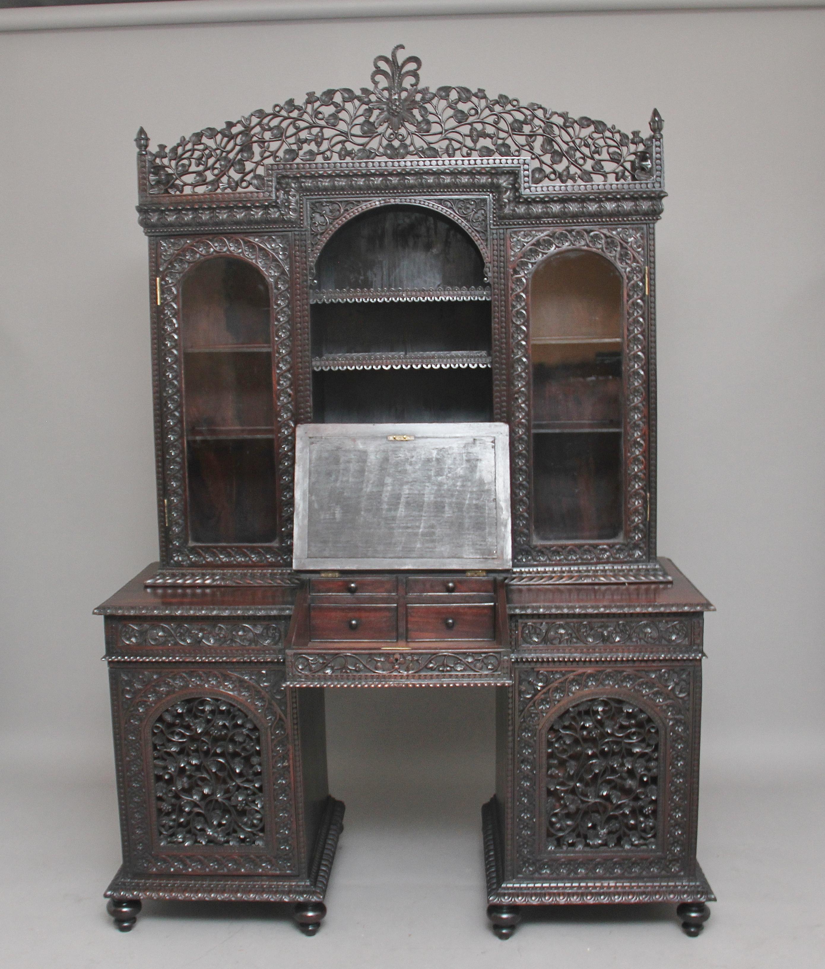 19th century Anglo-Indian hardwood bookcase desk, the base having two pedestals on turned feet, each pedestal with a heavily carved and pierced fret door which opens to reveal three drawers, above the pedestals is a section with two drawers flanking