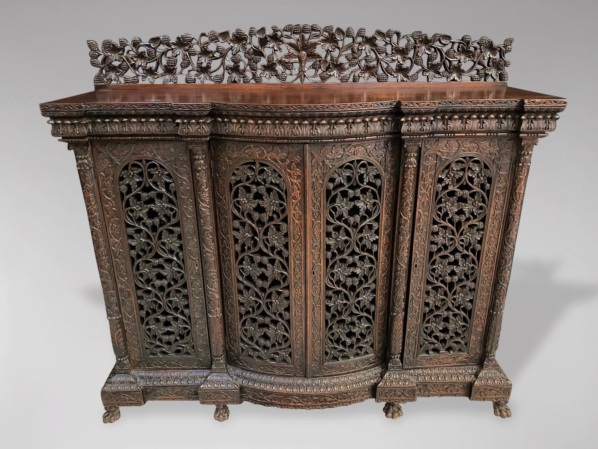 A mid 19th century, Victorian period, British colonial, Anglo-Indian carved and pierced serpentine sideboard in solid rosewood. The front is mounted with four hinged doors with dome shaped panelled doors enclosing shelves. All resting on six small,