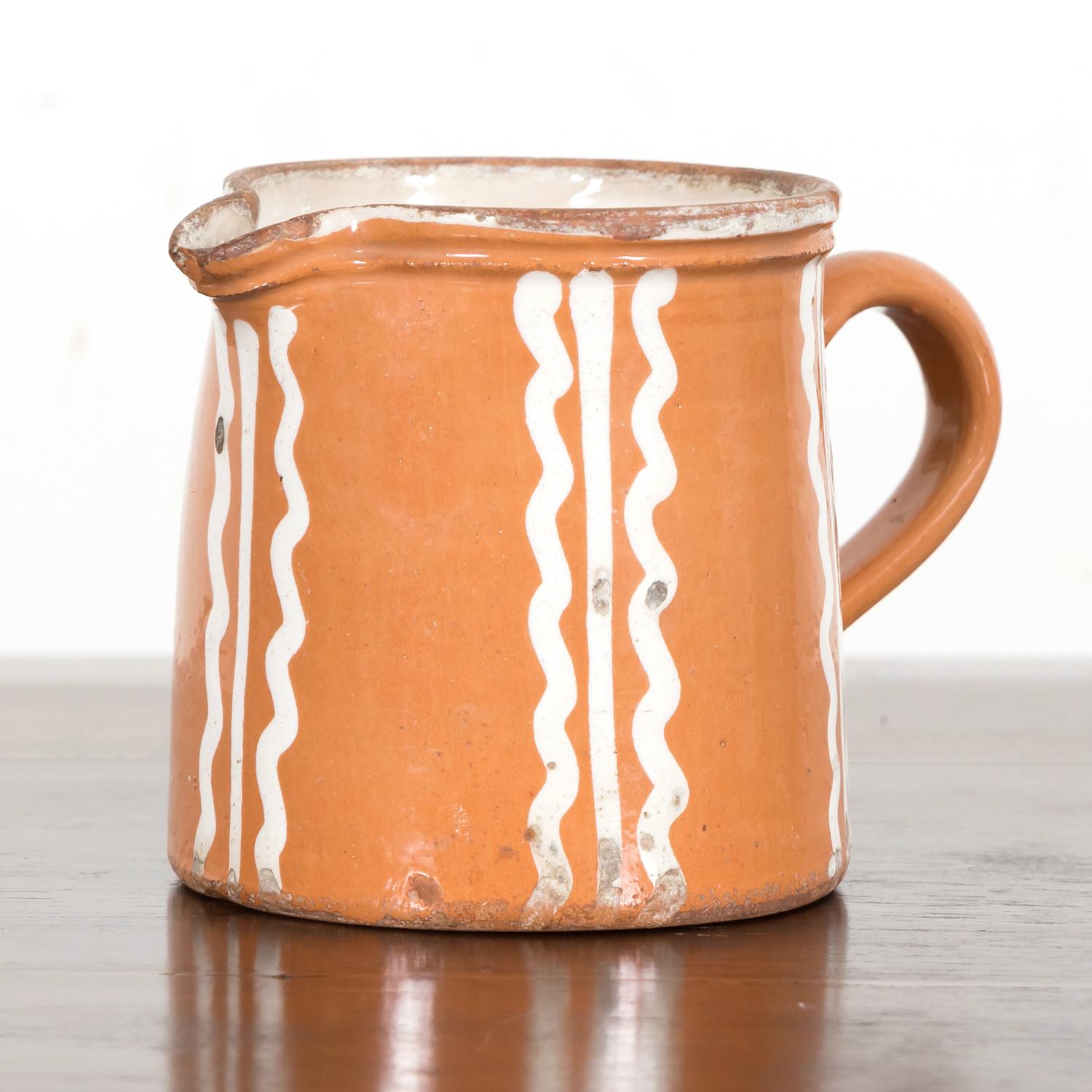 A beautiful 19th century French terracotta jaspé ware pottery pitcher, handcrafted near Annecy, in the Savoie region of France, circa 1870s. Having a large handle and small spout with burnt orange exterior glaze featuring hand painted cream colored