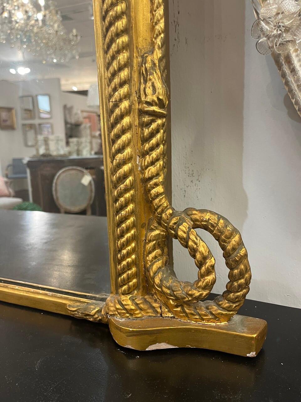 Este exquisito espejo antiguo de madera dorada presenta un cautivador borde con motivo de cuerda, que añade un toque de elegancia a cualquier espacio. Elaborados adornos adornan la montura, mostrando una artesanía intrincada y un encanto atemporal.