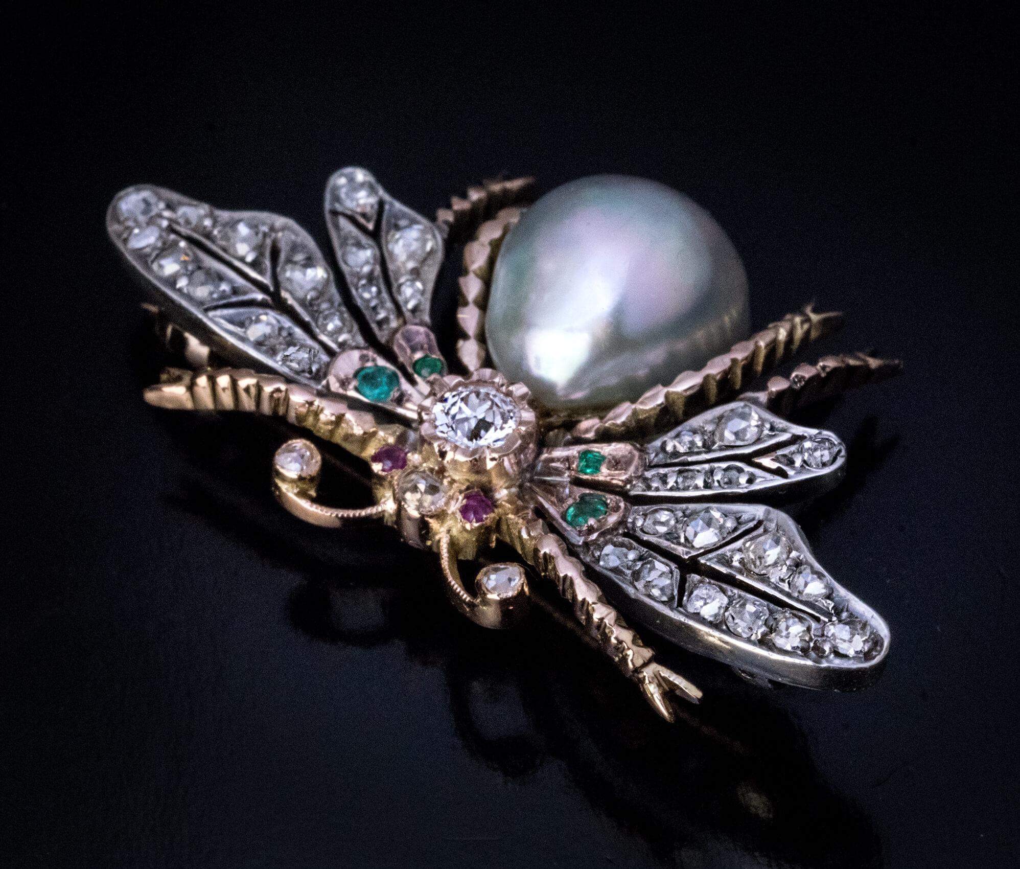 Circa 1890  The brooch is crafted in 14K gold and silver. A large pear shape pearl forms the body. The wings are embellished with old rose cut diamonds and emeralds. The head is highlighted by a sparkling old mine cut diamond. The eyes are set with