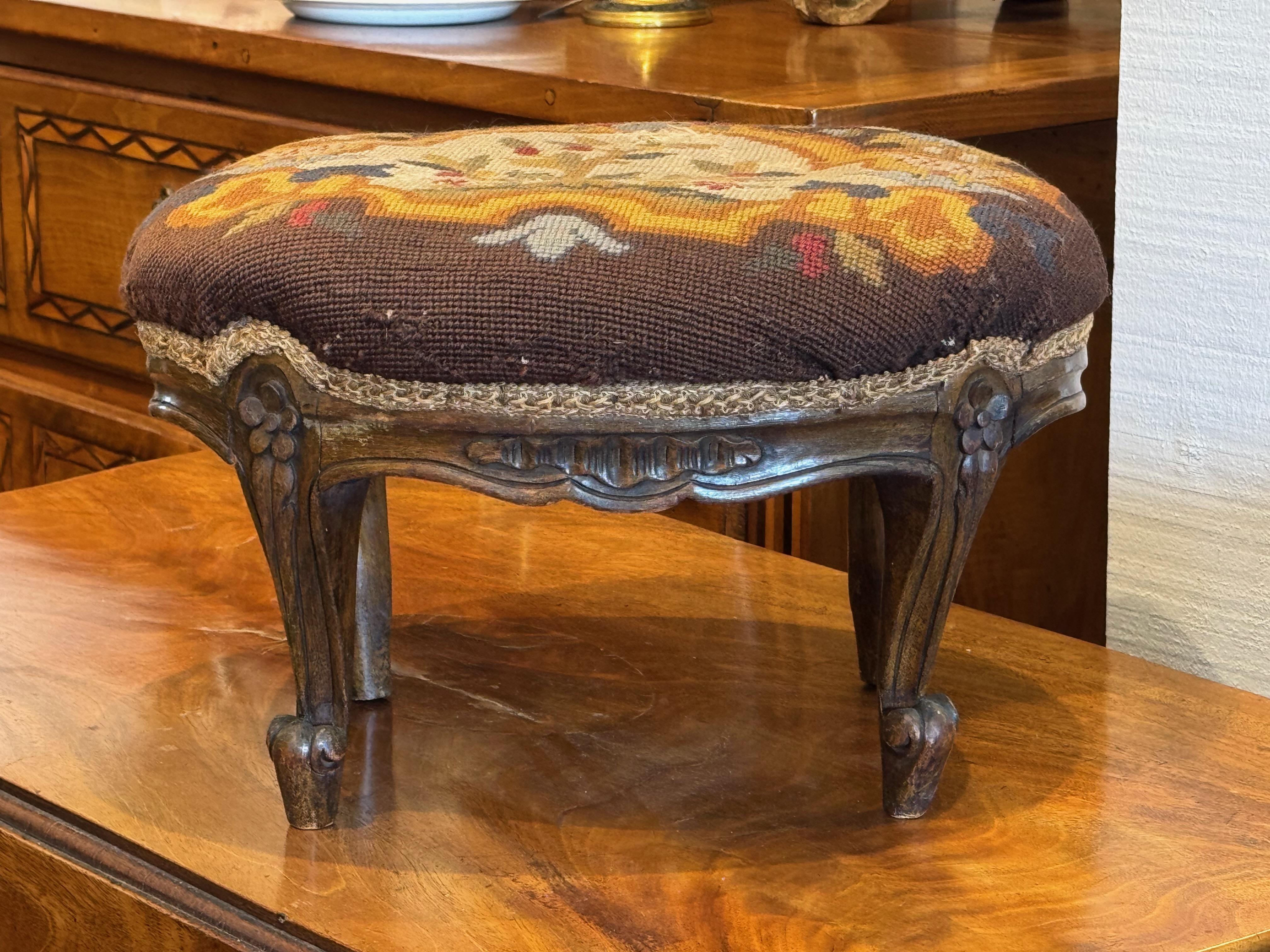 Charmant pouf ancien dont le dessus est orné d'un motif floral cousu à la main dans des tons chauds. La base en bois sculpté présente des accents floraux et des pieds en cabriole, reflétant une influence européenne classique. Une pièce décorative et