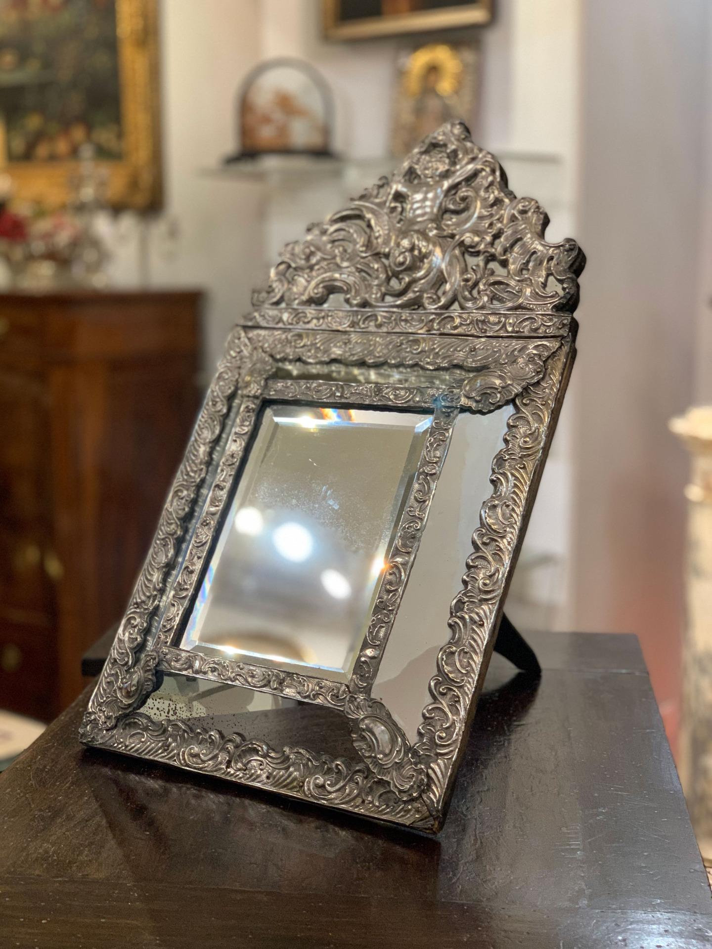 Elegant toilette table mirror in Renaissance style, in hand-crafted brass with floral decorations with the image of Cupid centrally. The mirror, which has a folding and disappearing pedestal, can also be placed on the wall, since its pedestal does