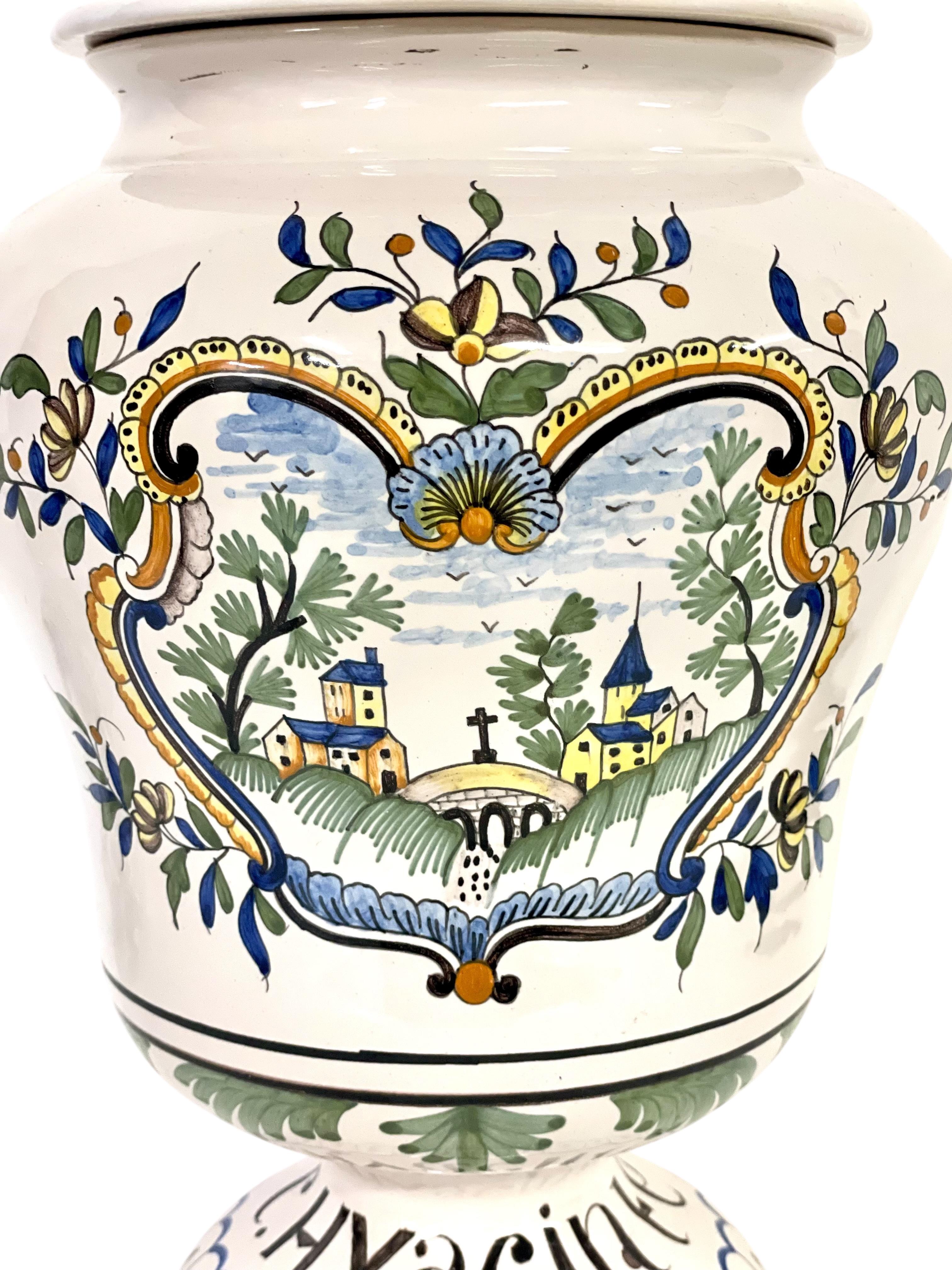 Questo favoloso vaso da farmacia francese d'epoca, con il suo coperchio a chiusura ermetica e la sua base a piedistallo, è stato splendidamente dipinto a mano con una scena rurale e provenzale abbellita da semplici fiori e foglie. Sulla base è