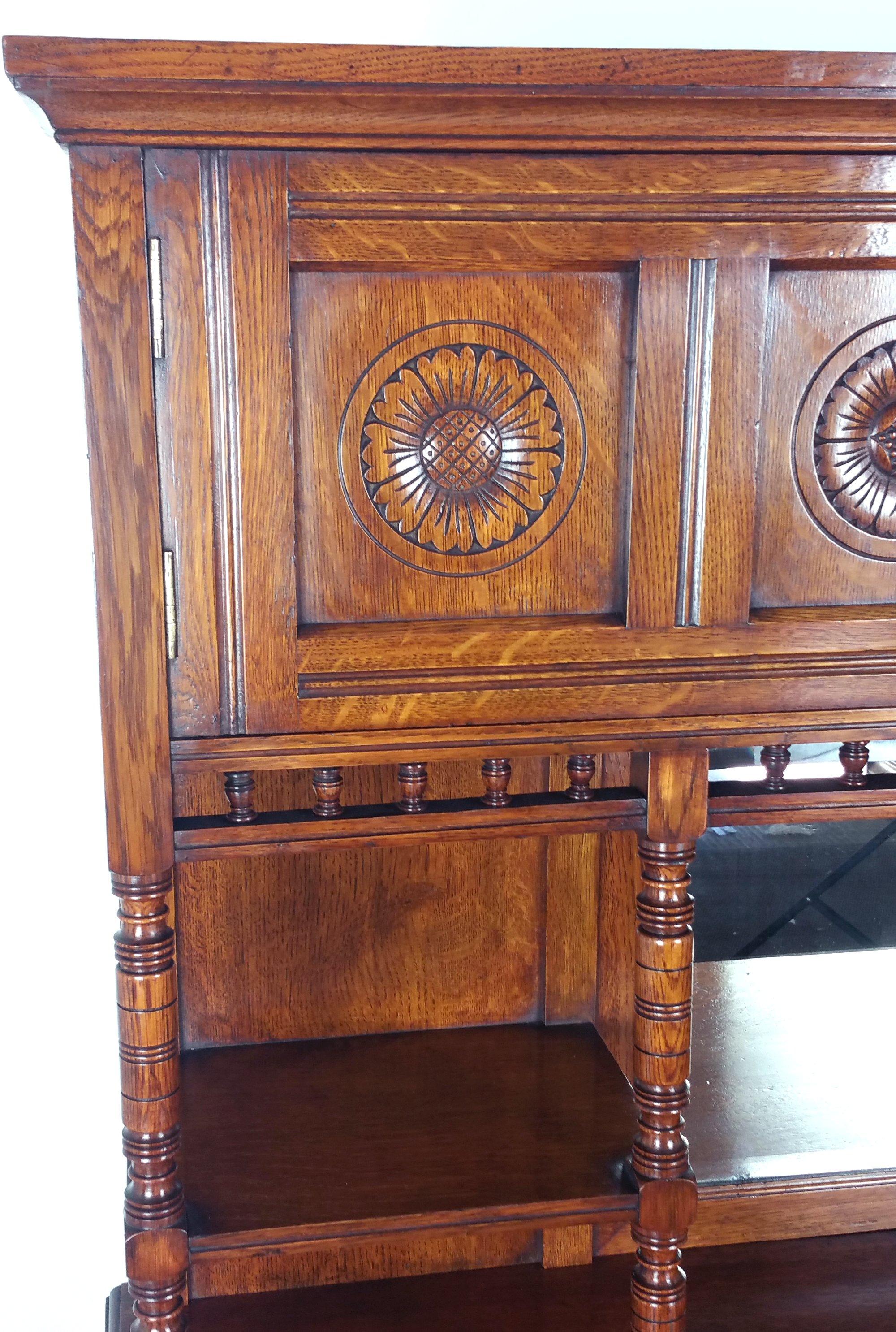 This beautiful 19th century. English oak Arts & Crafts sideboard features a central bevelled mirror with shelf units on either side with turned support detailing. The top has 2 doors with stylised sunflower designs on each of the four panels. The