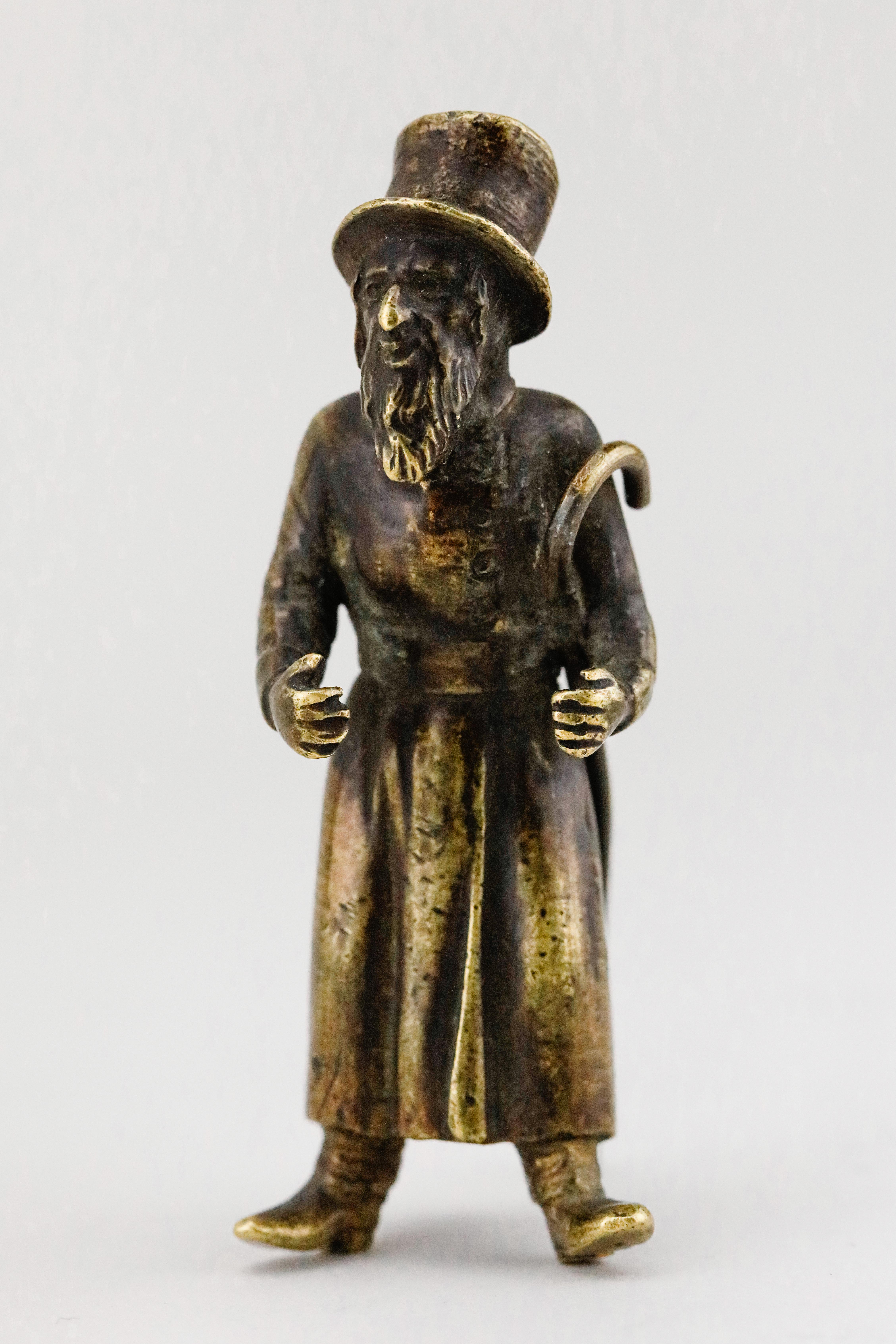 Cast bronze figurine of a Jewish man in a top hat, ankle-length overcoat, and heeled boots. He holds a crook umbrella handle on his left arm. He has thick, grooved sidelocks, moustache, and long beard with large, deep set eyes under thick brows, and