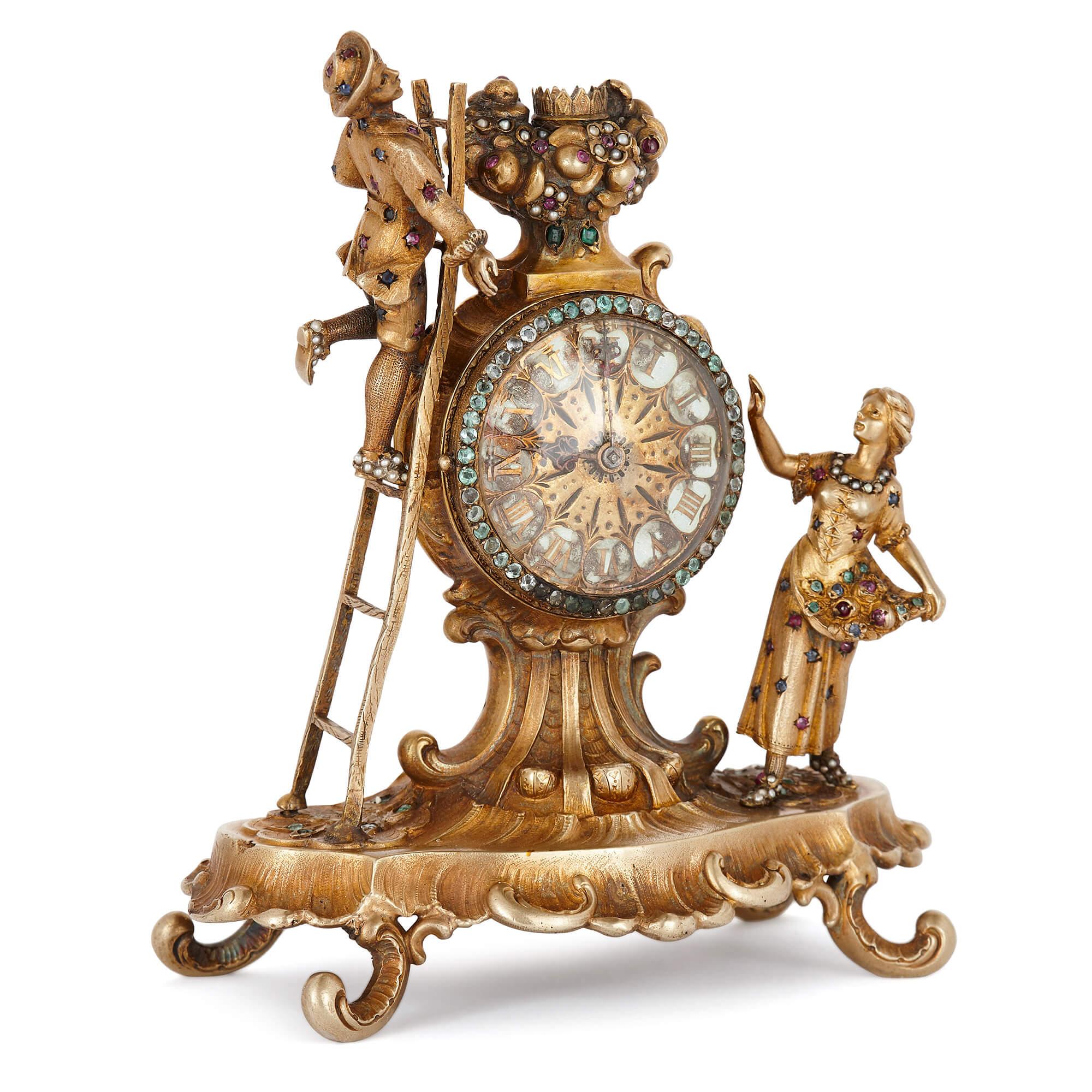 This jewel-encrusted, silver-gilt antique table clock is an exquisite piece of Viennese craftsmanship. It comprises of a scalloped base, which is raised up on four C-scroll feet, and topped by a crowned circular enameled dial. Flanking the dial are