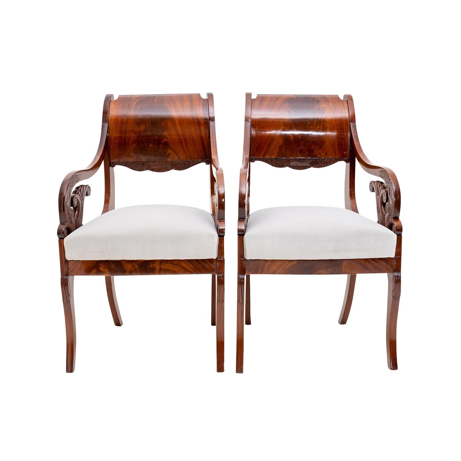 A dark-brown, antique Baltic Biedermeier pair of armchairs made of handcrafted polished Mahogany, in good condition. The particularized veneered carved side chairs have a wide arched backrest withs slim arms, standing on four slightly curved wooden