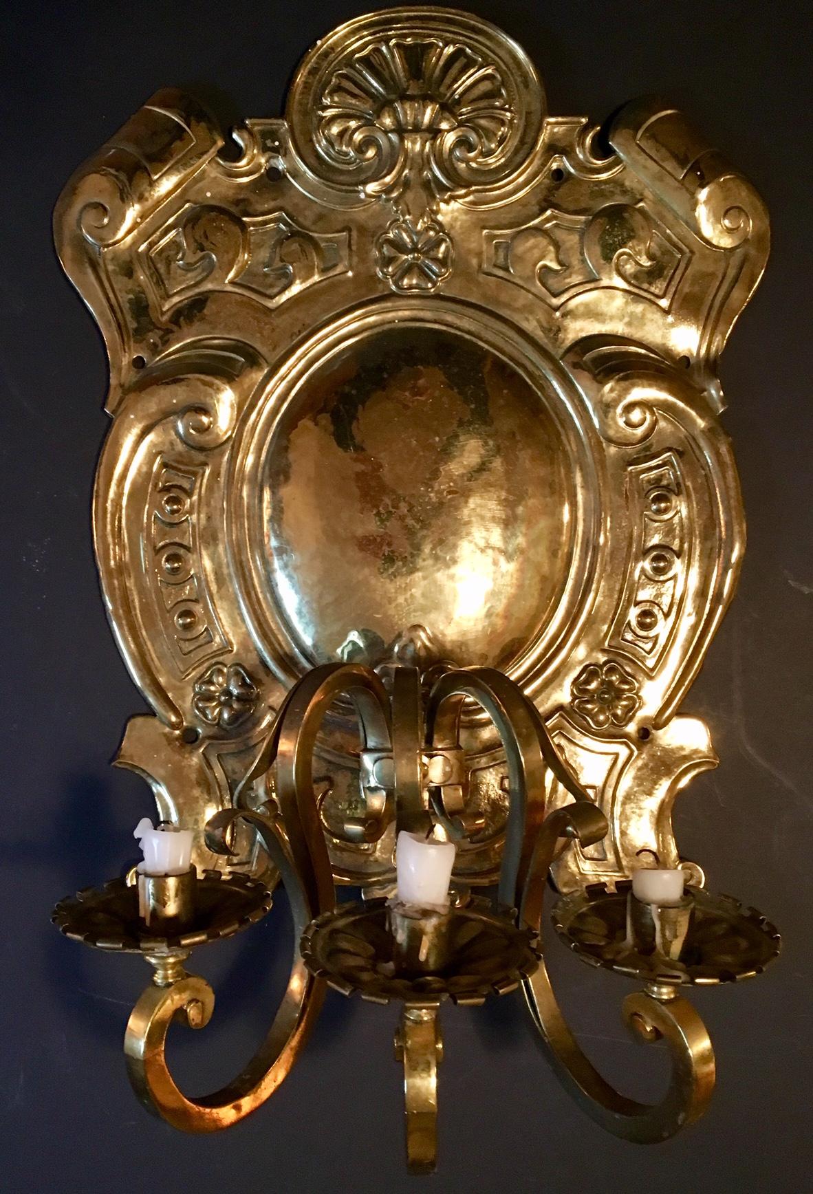 This is an impressively large and ornate antique repousse brass three-light sconce. The backplate is crest shaped with a convex center embossed with a shell and scroll design. The Dutch Baroque shaped candle arms are complete with cups and bobeches.