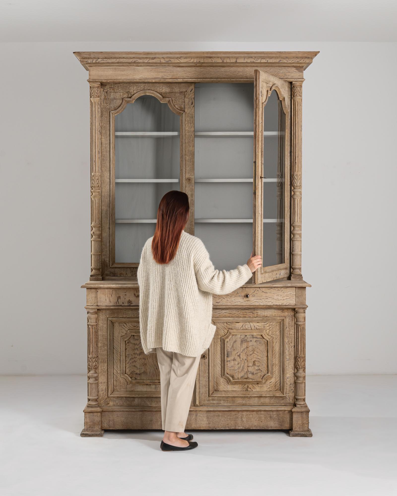 Made in the 19th century in Belgium, this oak vitrine impresses with its robust complexion and the richness of its decorative elements. The pronounced molded cornice lends a neoclassical touch, enhanced by gracefully carved arches, and columns. With