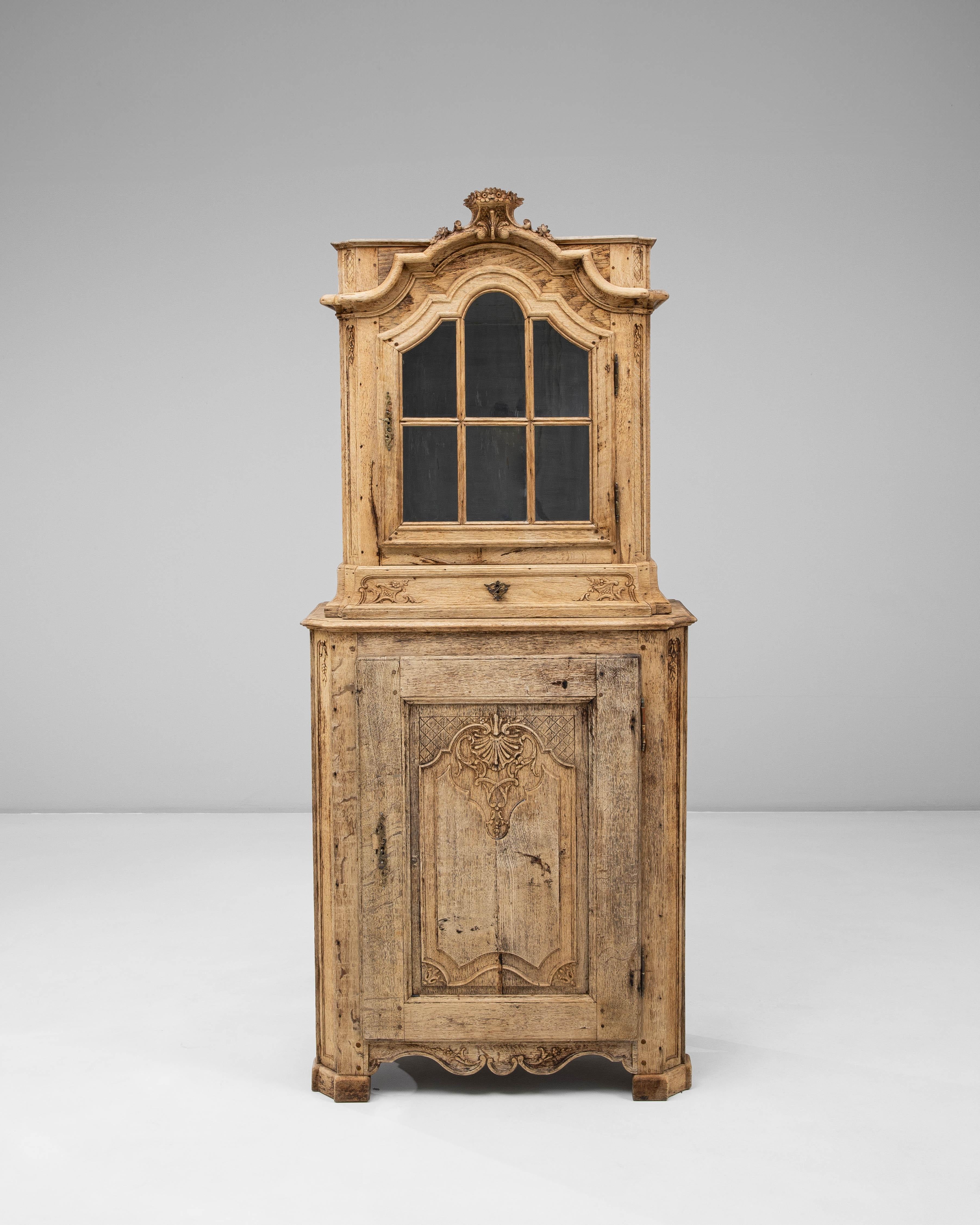 Questo mobile belga in quercia Luigi XV del XIX secolo mostra la graziosa curvatura e i dettagli ornamentali caratteristici dell'epoca, espressi attraverso l'elegante piano ad arco, lo stemma intagliato e l'anta sagomata con pannelli di vetro. La