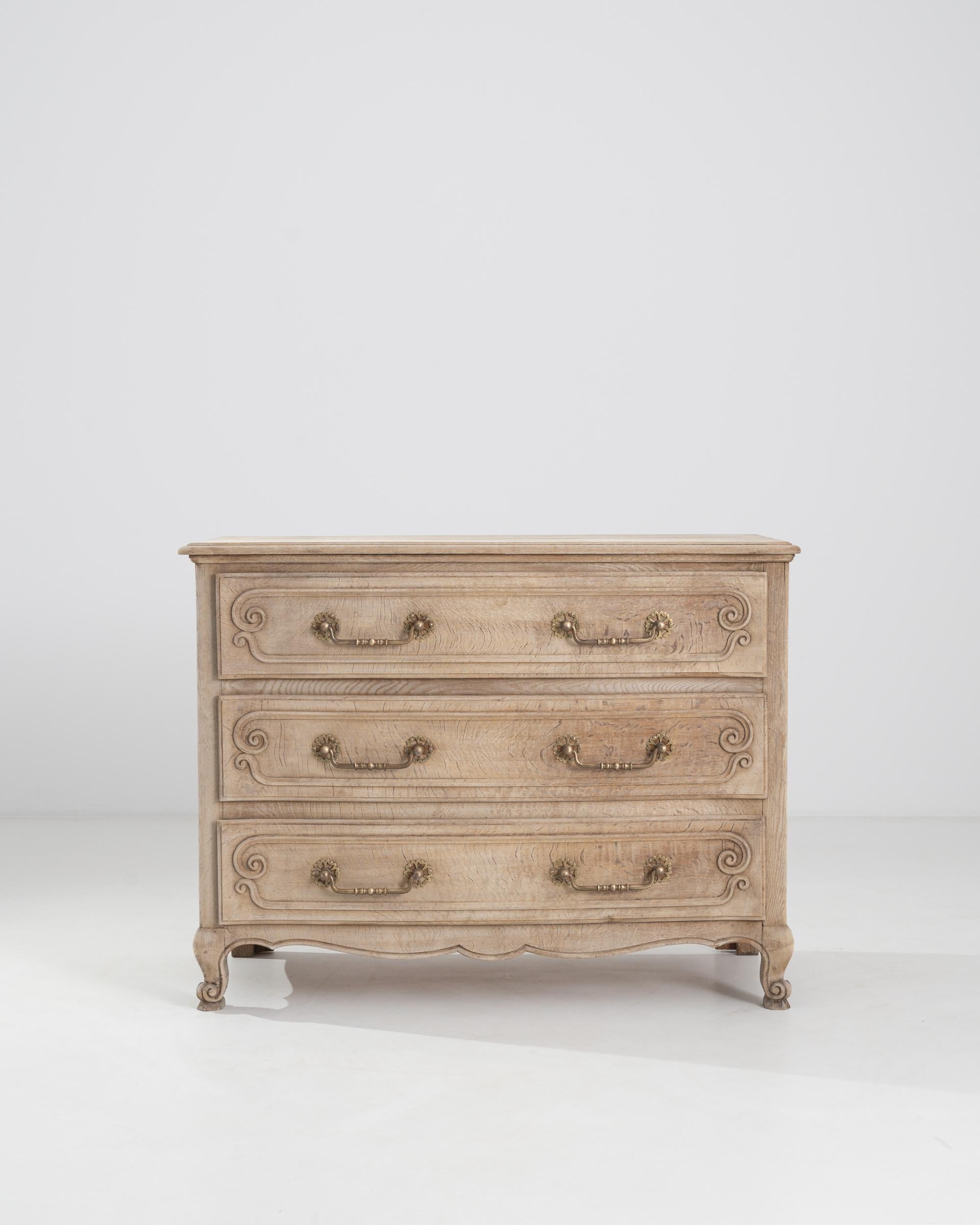 A bleached oak chest of drawers from 20th century Belgium. This chest wields three spacious drawers, a flowing wave runs along its bottom edge, flanked by carefully carved scroll motifs along its front-facing feet. A sensitive bleaching process has