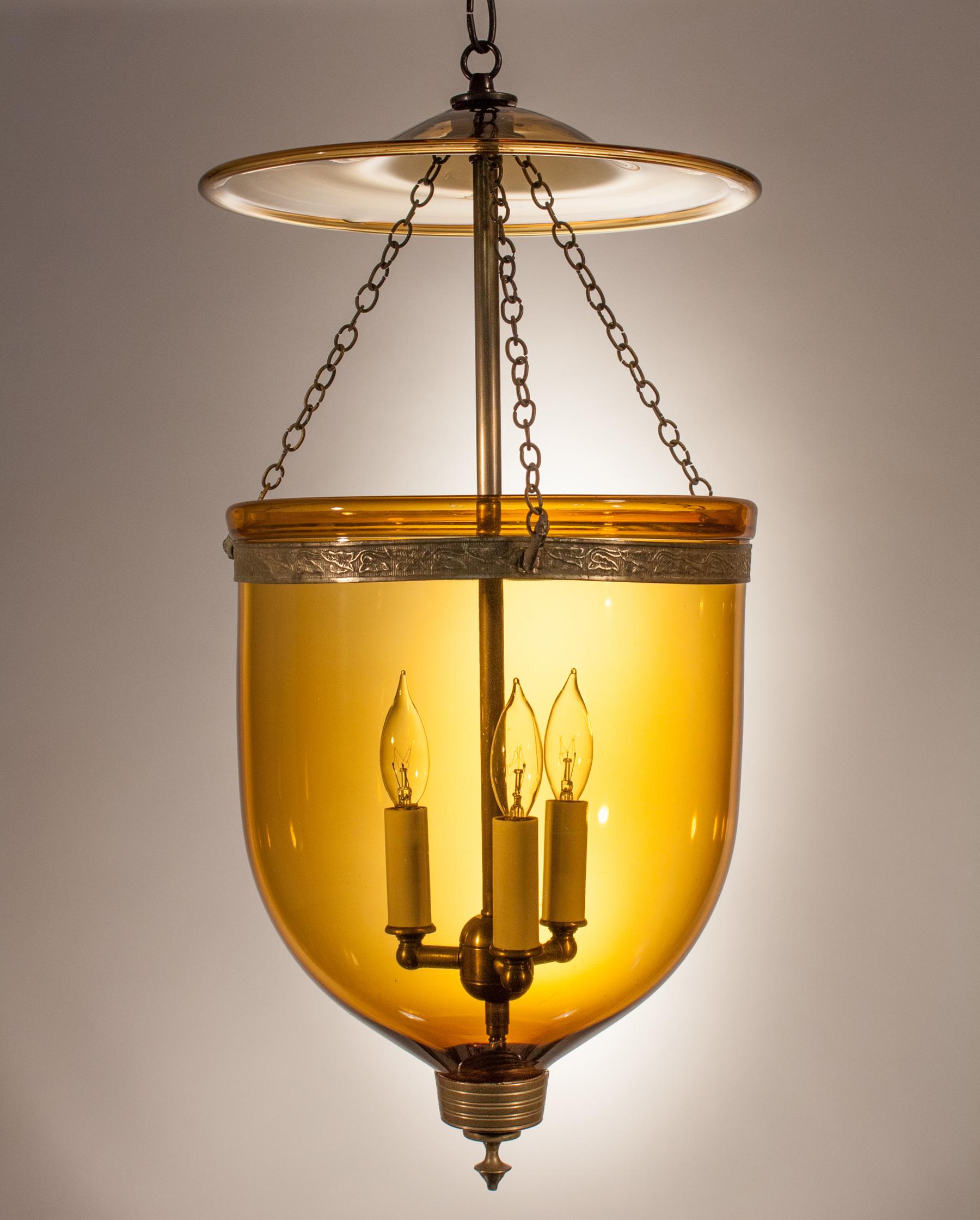 A rich amber-colored hand blown glass bell jar lantern with its original embossed brass band, finial and glass smoke bell. Chains are replaced, but originals are available. At first used with candles, this circa 1880 lantern has been newly re-wired