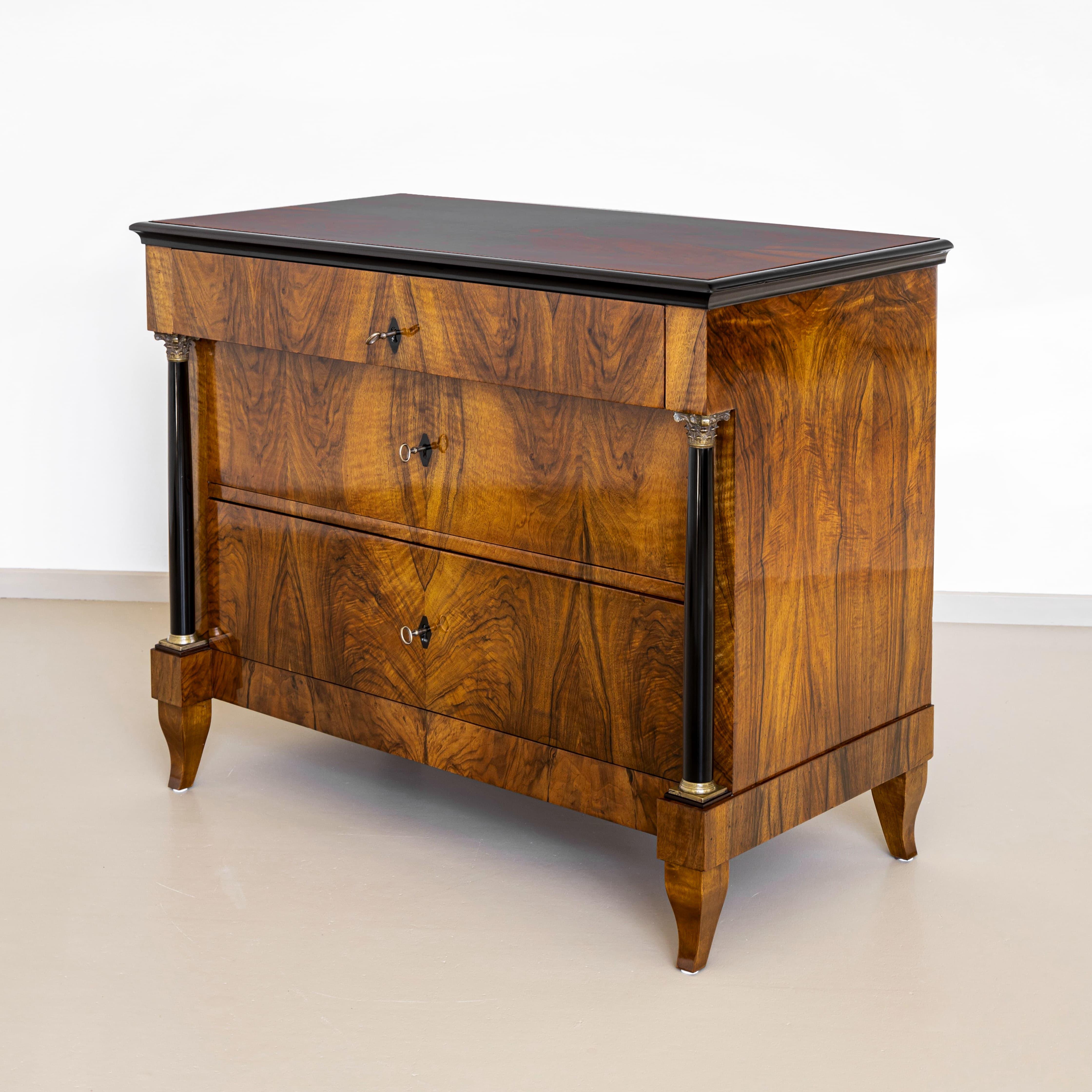Cómoda Biedermeier sobre patas en forma de S con columnas ebonizadas, bases y capiteles de latón. El borde perfilado circundante también está ebonizado. La cómoda ofrece mucho espacio de almacenamiento con tres cajones. La cómoda está chapada en