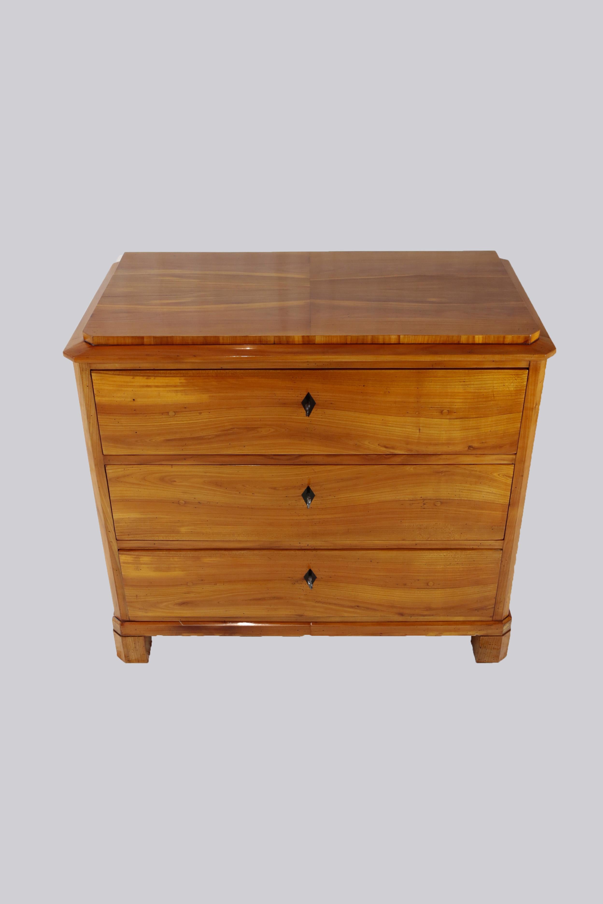 The Biedermeier chest of drawers with 3 drawers, designed in a straight line, was created around 1820-1830. The straight lines of the chest of drawers are accentuated by the stepped top and the transverse veneer on the drawers. Each of the drawers