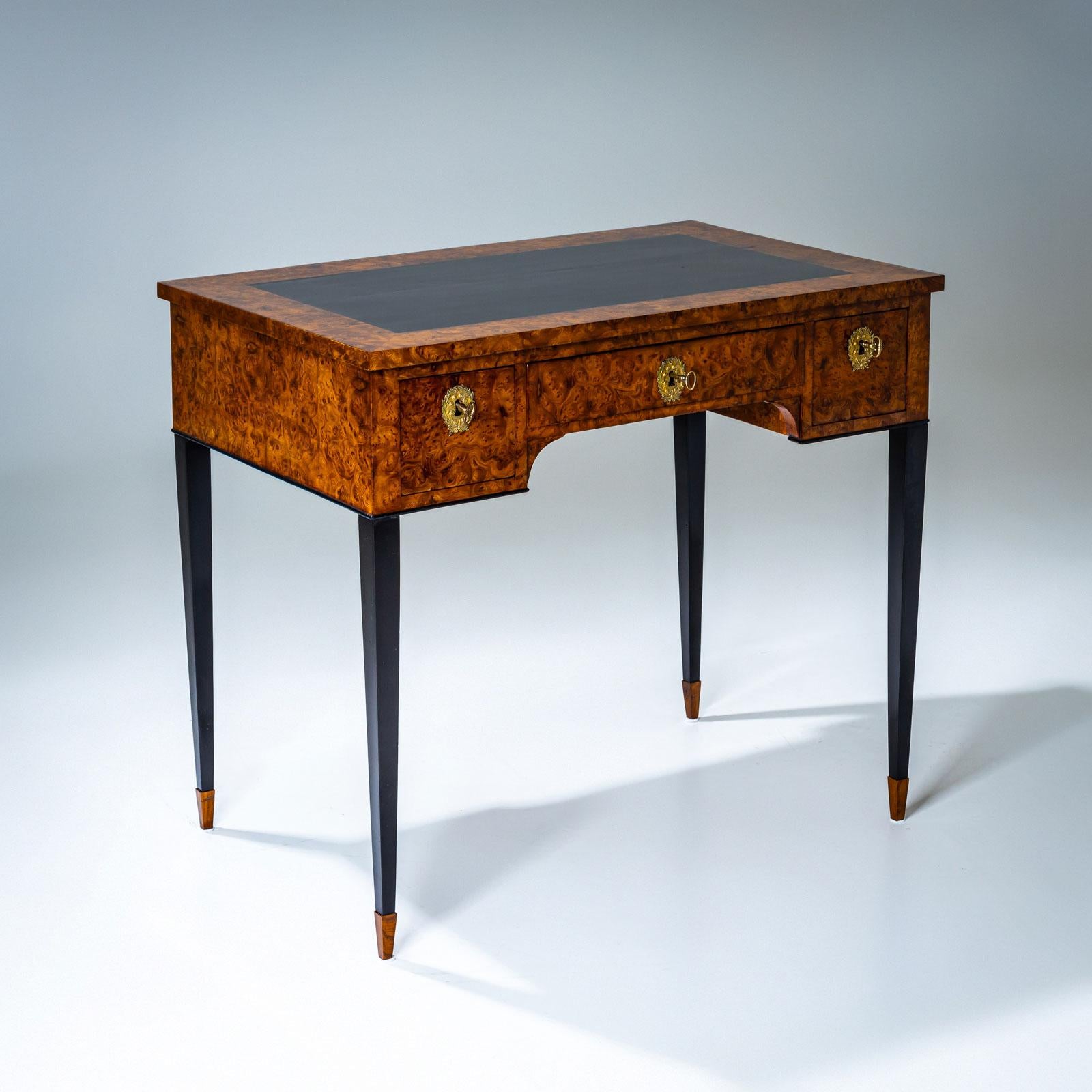 Small desk with a burlwood veneer and black lacquered writing surface. The desk stands on ebonized tapering legs and offers sufficient storage space with three drawers. The fittings are made of gilded bronze. The legs were replaced at a later date.