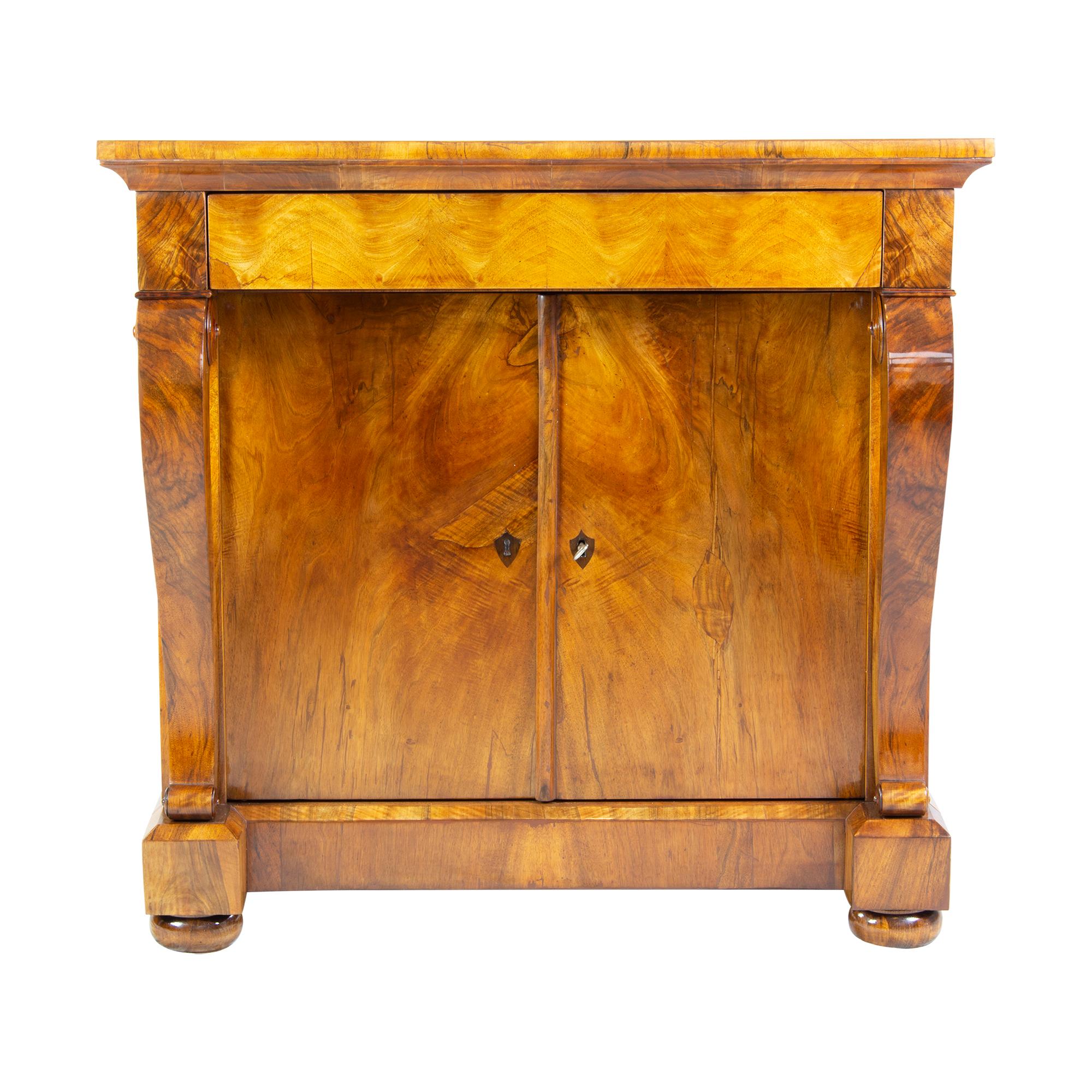 The furniture is a console half cabinet from the Biedermeier period. The half cabinet is walnut veneered on a spruce corpus. There is a drawer under the top and under it are the two doors, which are lockable together. The corpus stands on ball feet