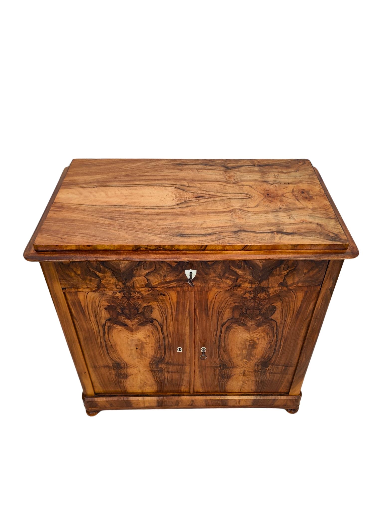This charming half cabinet dates back to the late Biedermeier period around 1830 and showcases timeless elegance. Crafted with a solid spruce body, it is adorned with a rich walnut veneer that highlights the warm, natural grain of the wood. The