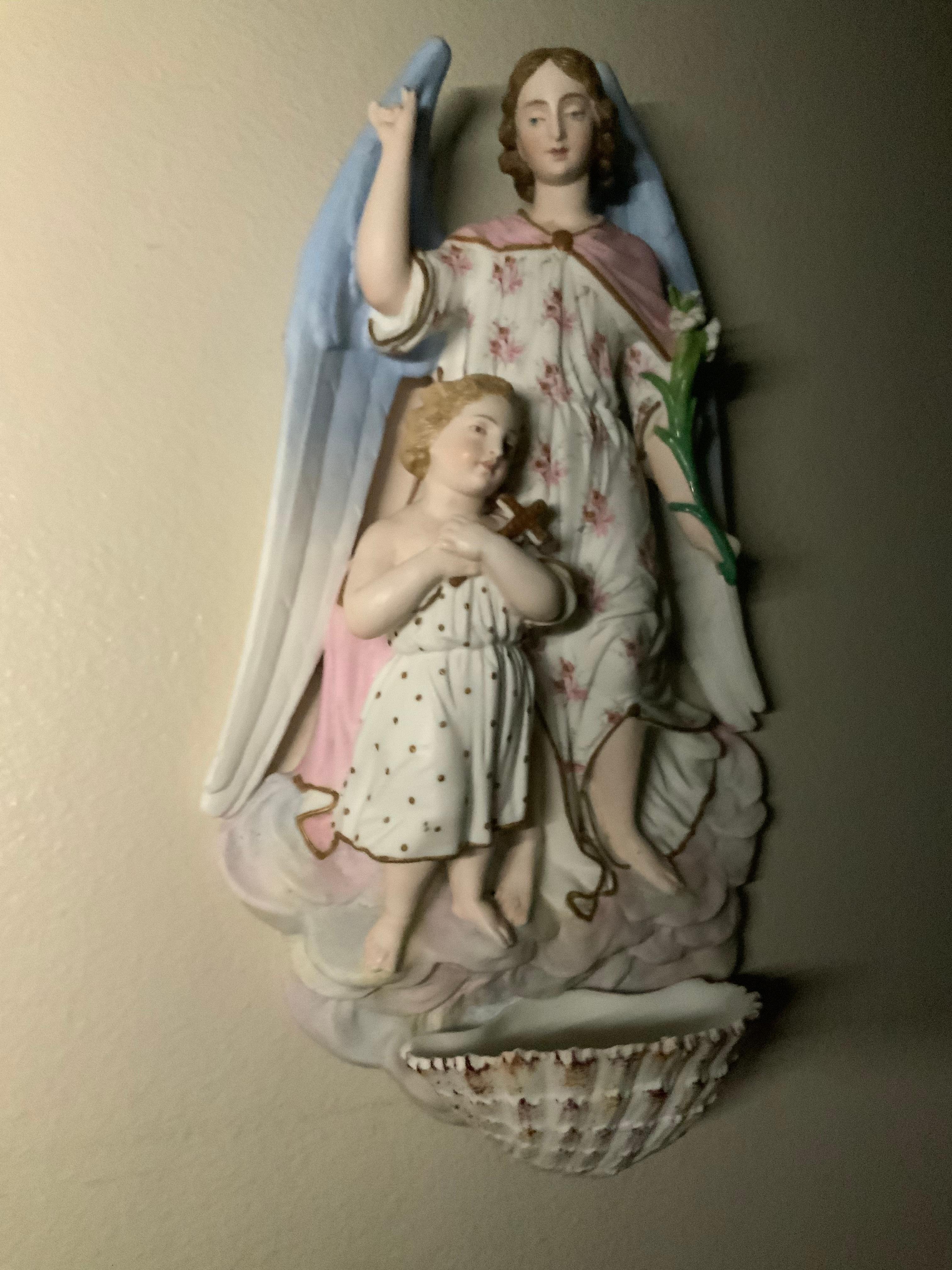 This is a hand painted bisque porcelain sculpture of a large winged guardian angel behind a child. The angel is dressed with a white robe printed with pink flowers and a pink cape and is holding a branch of Calla lilies in the left hand. The large